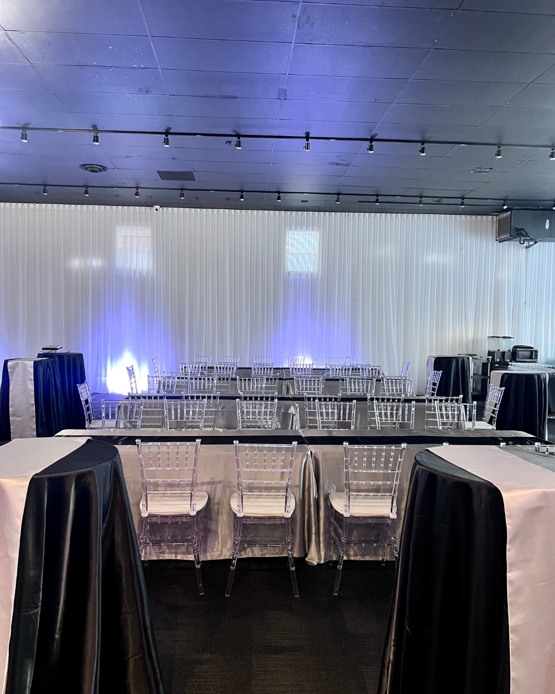 Empty conference or banquet room with tables covered in black and white tablecloths, clear acrylic chairs, and a white curtain backdrop with blue lighting.