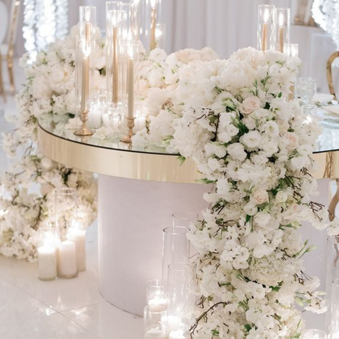 Elegant wedding table with white floral arrangements, candles, and a glass-topped table with a gold edge.