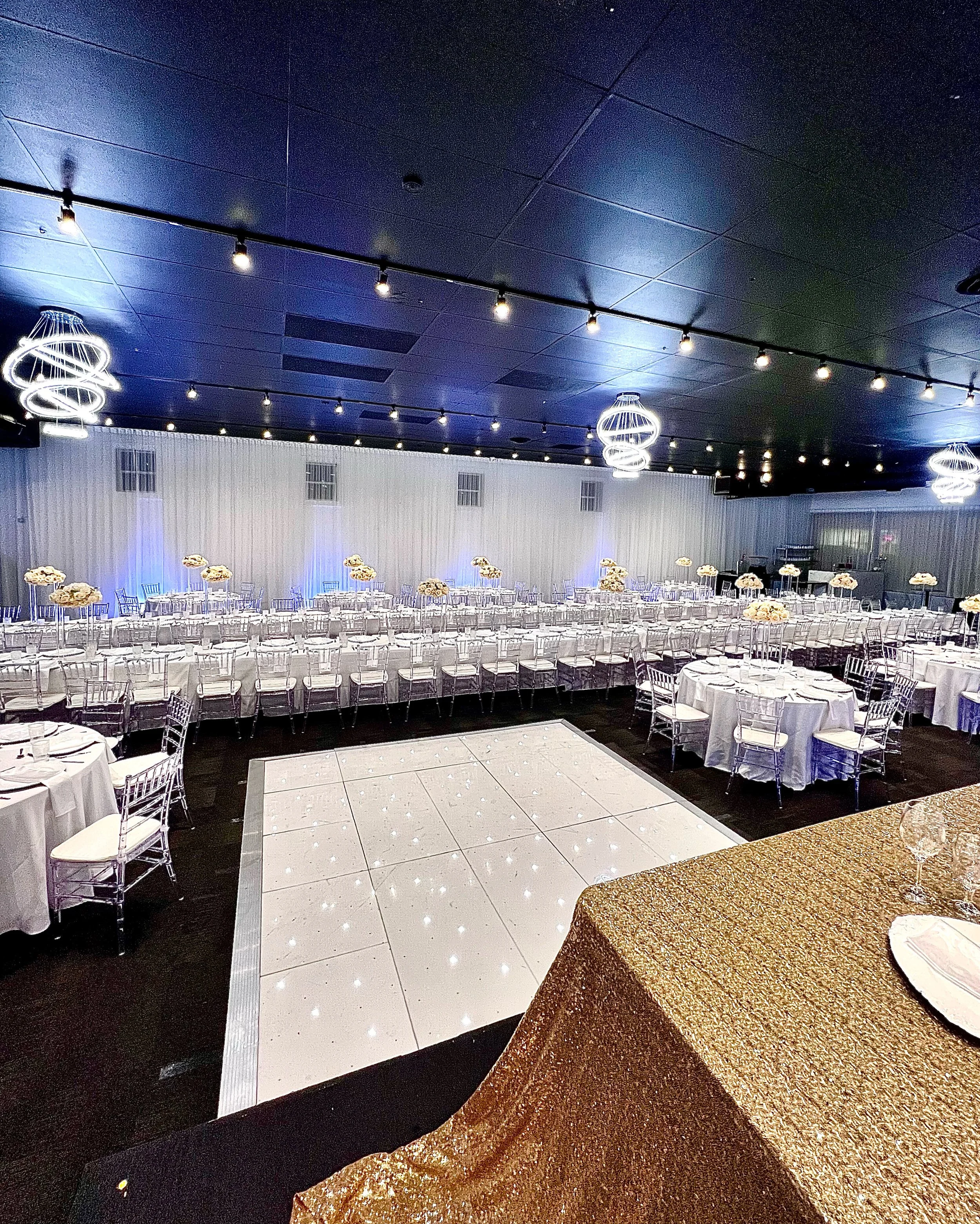 Elegant event hall decorated for a celebration with round tables covered in white tablecloths and floral centerpieces, clear chairs, a dance floor, modern spiral chandeliers, and soft blue lighting.