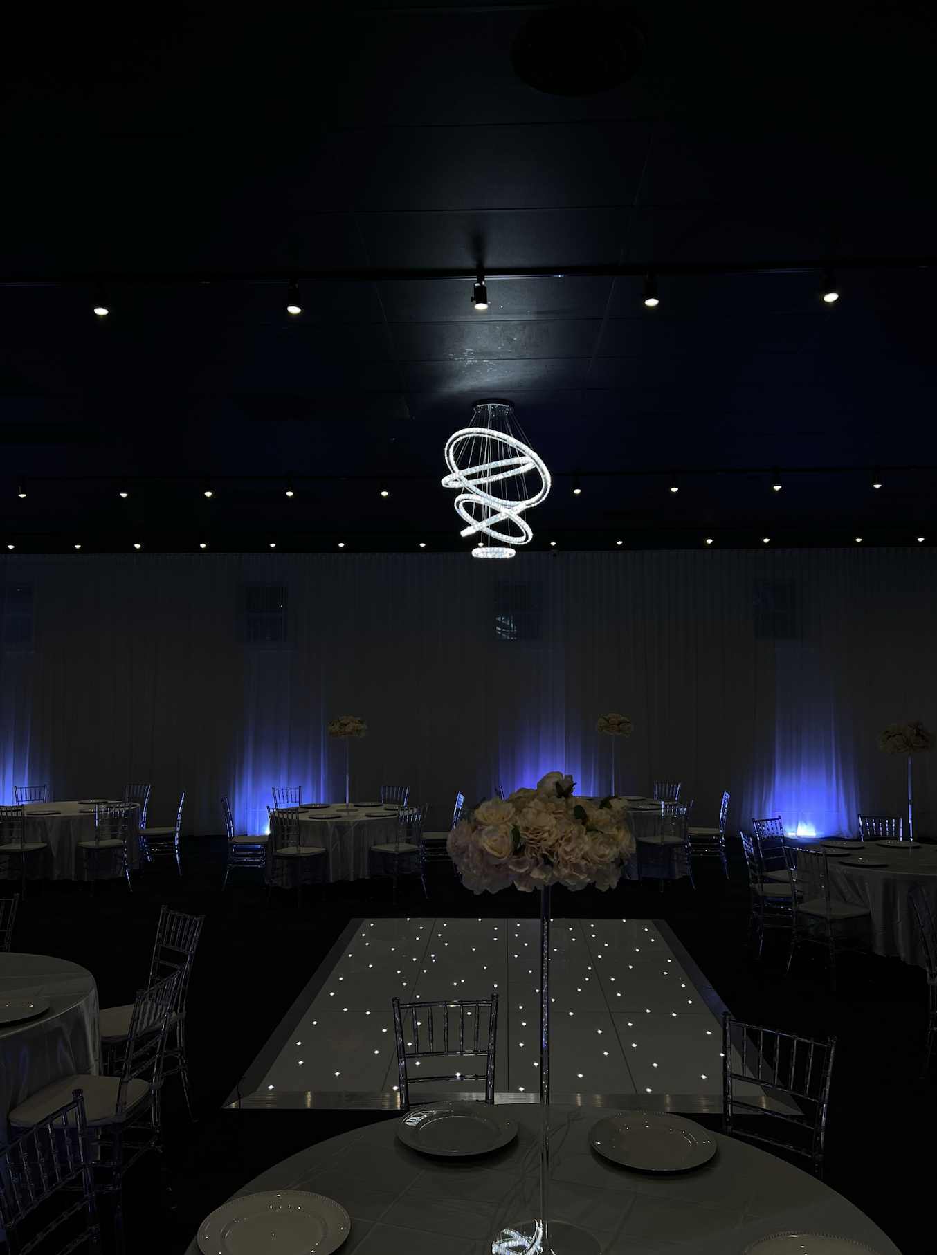 Elegant event space with round tables, floral centerpieces, and a illuminated dance floor. Modern spiral chandelier hanging from the dark ceiling with blue accent lighting along the walls.