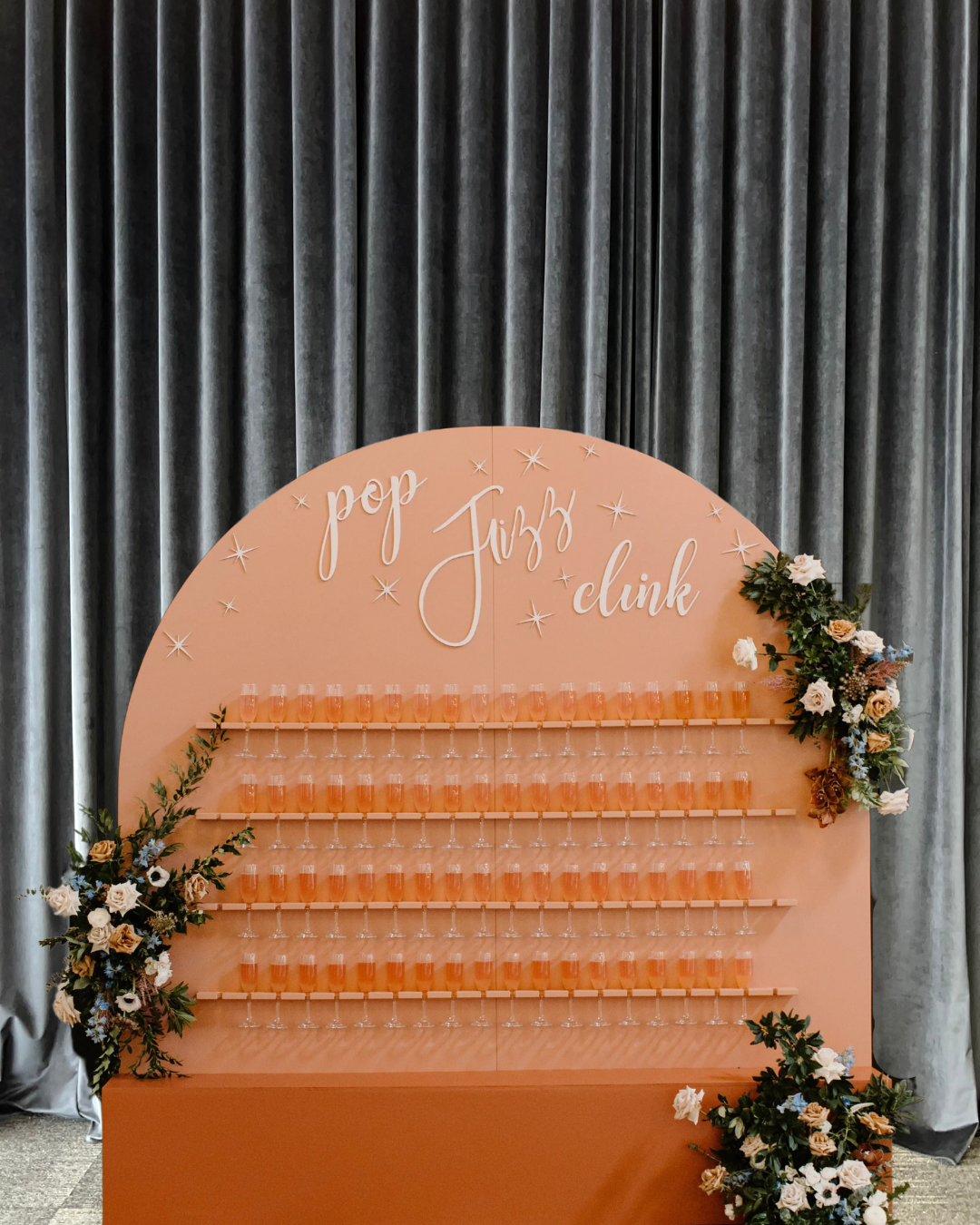 Pink display board with the words "pop Jazz clink" and rows of glasses filled with a pink drink, decorated with floral arrangements on the sides, set against a dark curtain background.