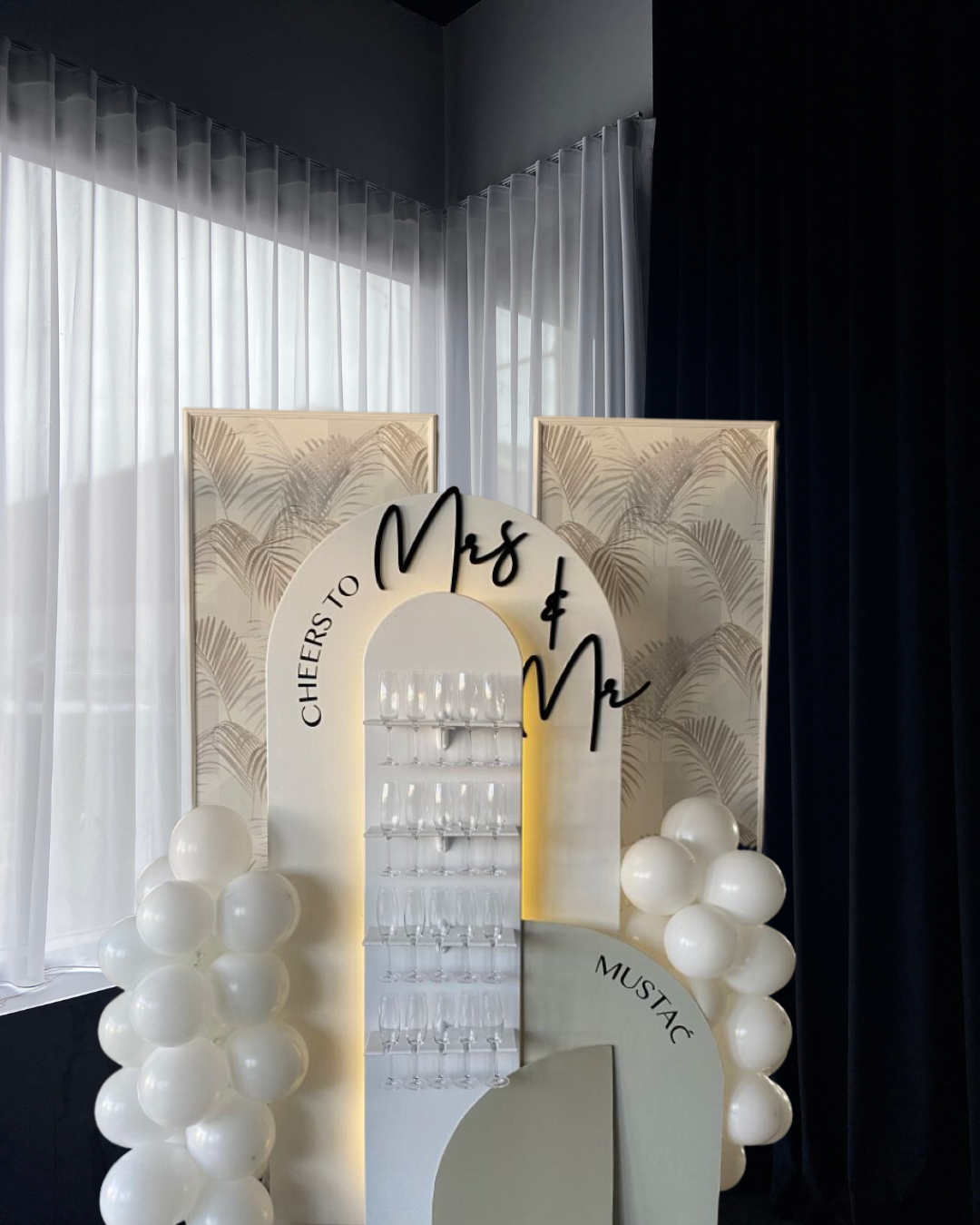 Wedding bar display with a Cheers to Mrs. and Mr sign, white balloons, and glasses on shelves, set against white and dark curtains.
