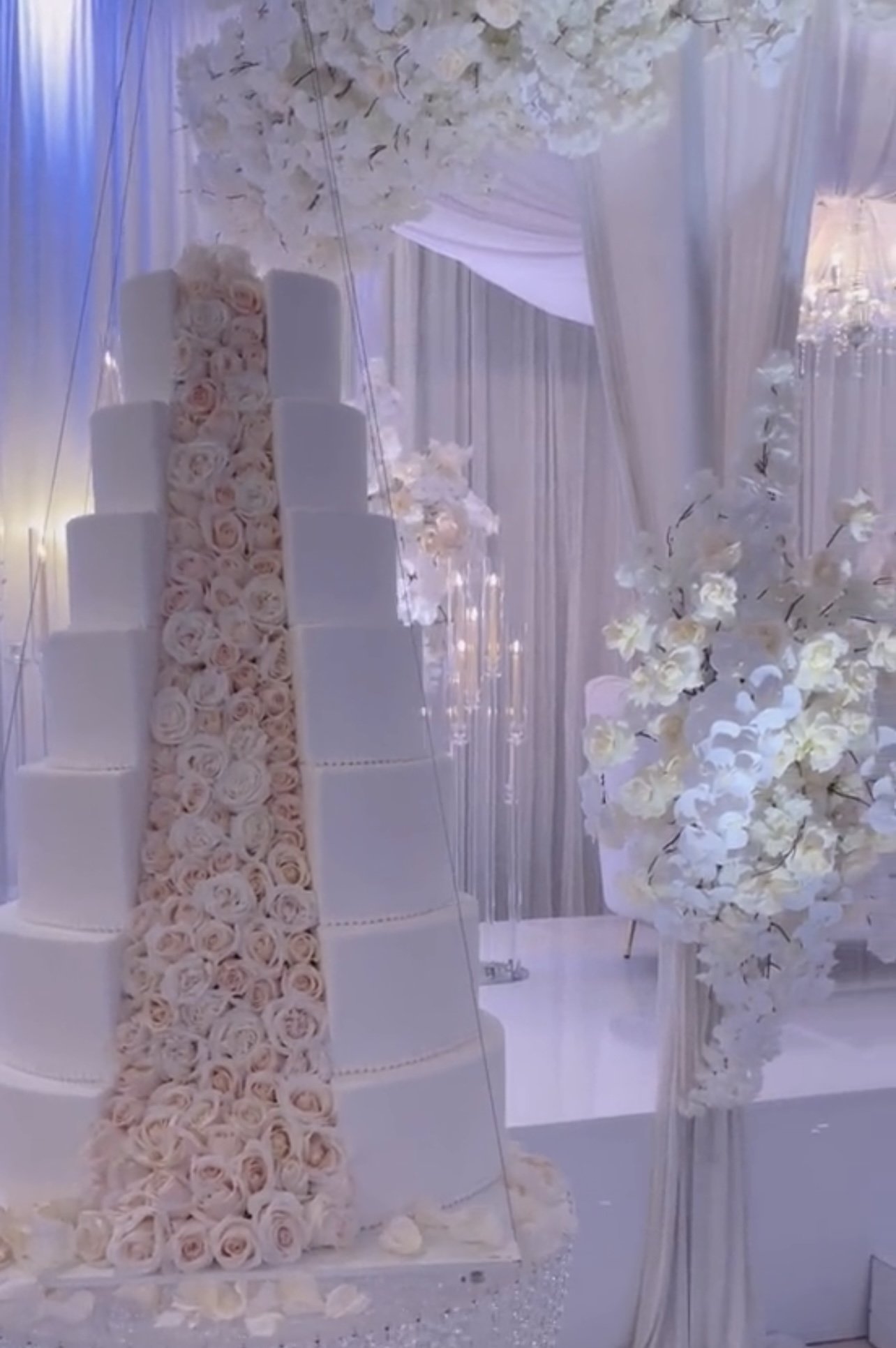 A wedding cake decorated with pink roses and white flowers, with a floral aisle runner and elegant drapery.