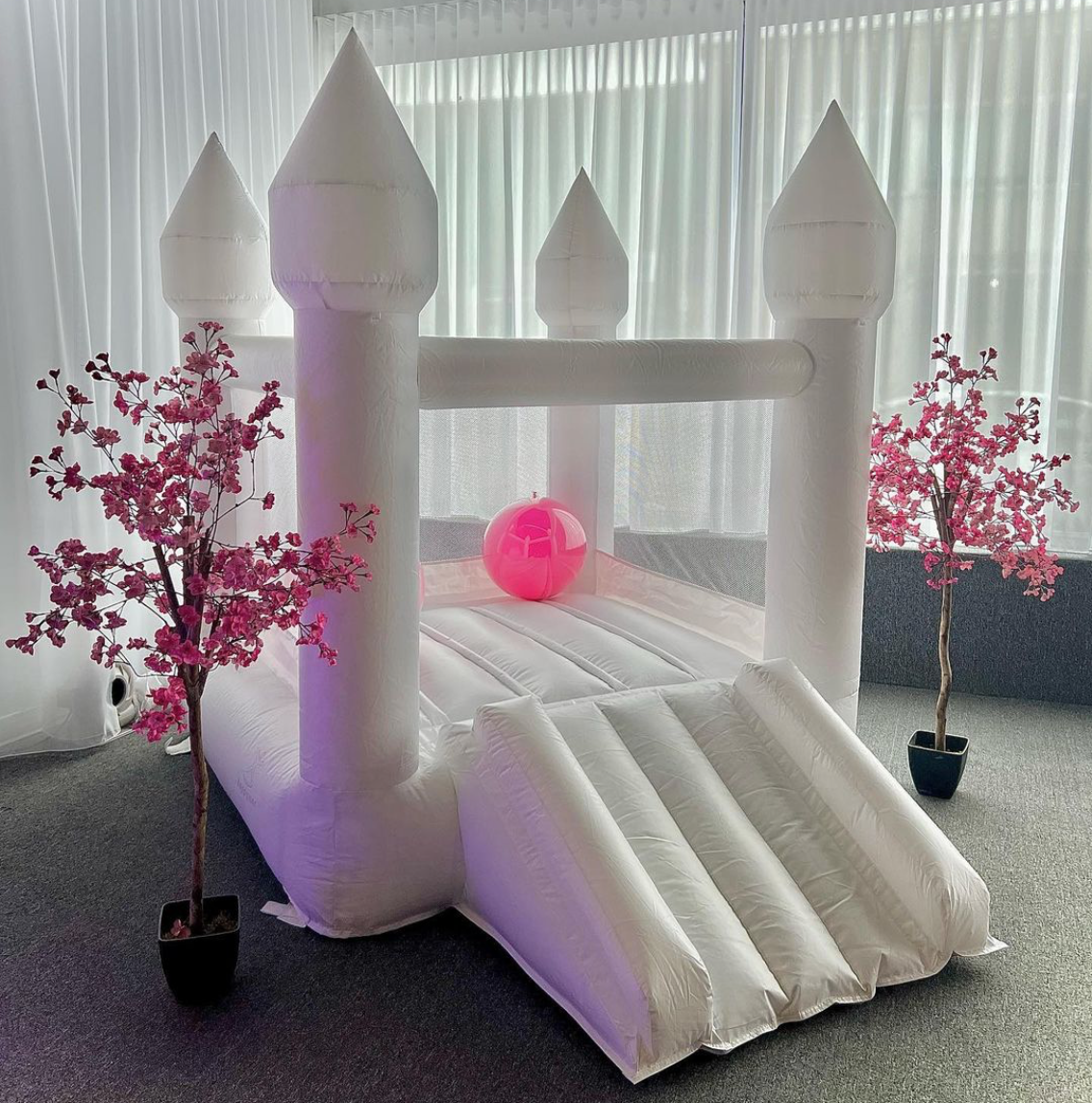 An indoor inflatable white castle bounce house with pink cherry blossom trees on either side and a pink ball inside, set up on gray carpet in front of large windows with sheer curtains.