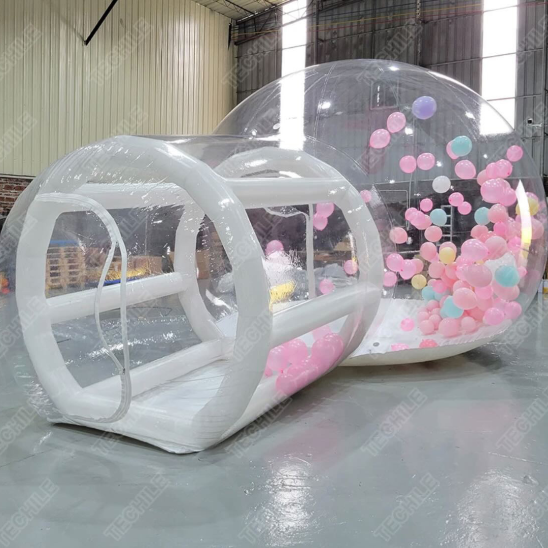 Large transparent inflatable bubble with colorful balloons inside, set up indoors with high ceilings and metal walls.