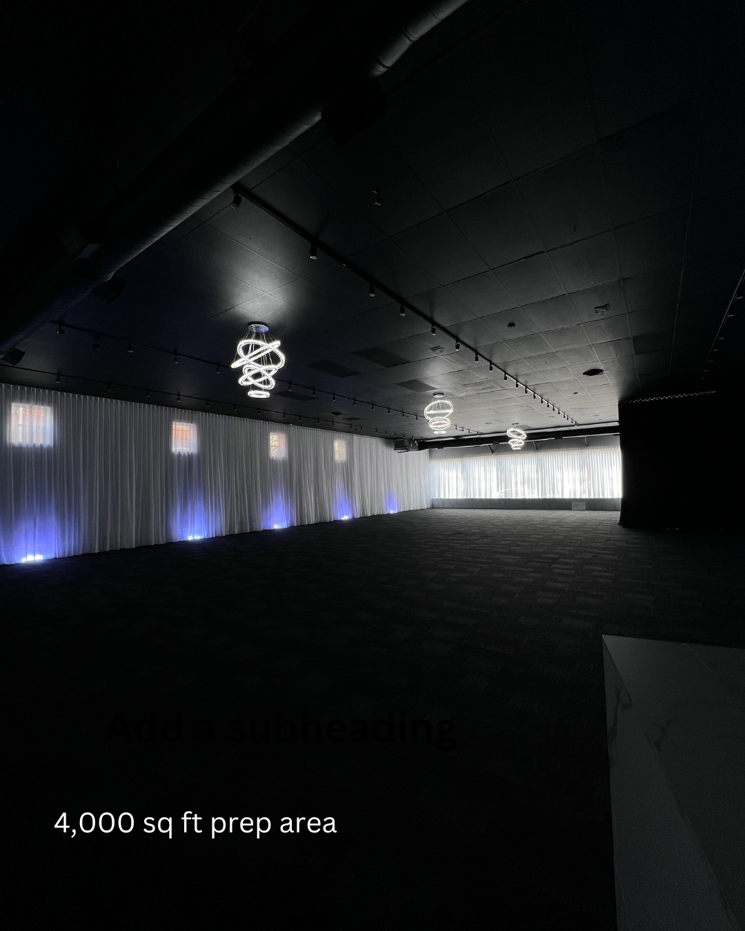 Empty 4,000 sq ft event preparation area with dark ceiling, modern spiral light fixtures, white curtain wall with blue accent lighting, and large windows at the far end.