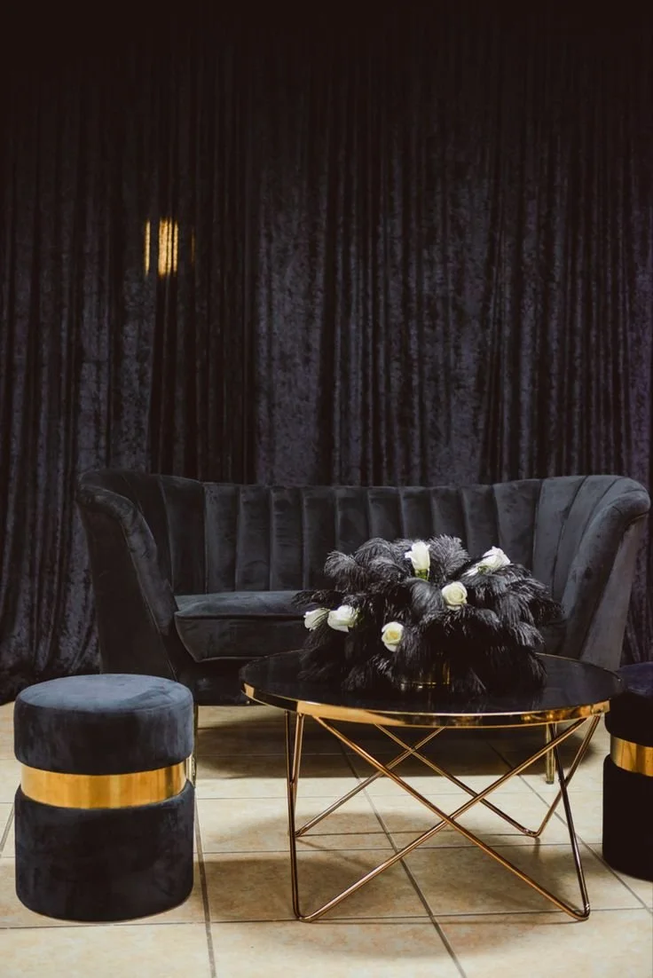 A black velvet settee with a matching round ottoman with gold accents, a black glass-topped coffee table with gold stands, and a floral arrangement of white roses and black feathers in a black vase, all against a dark velvet curtain background.