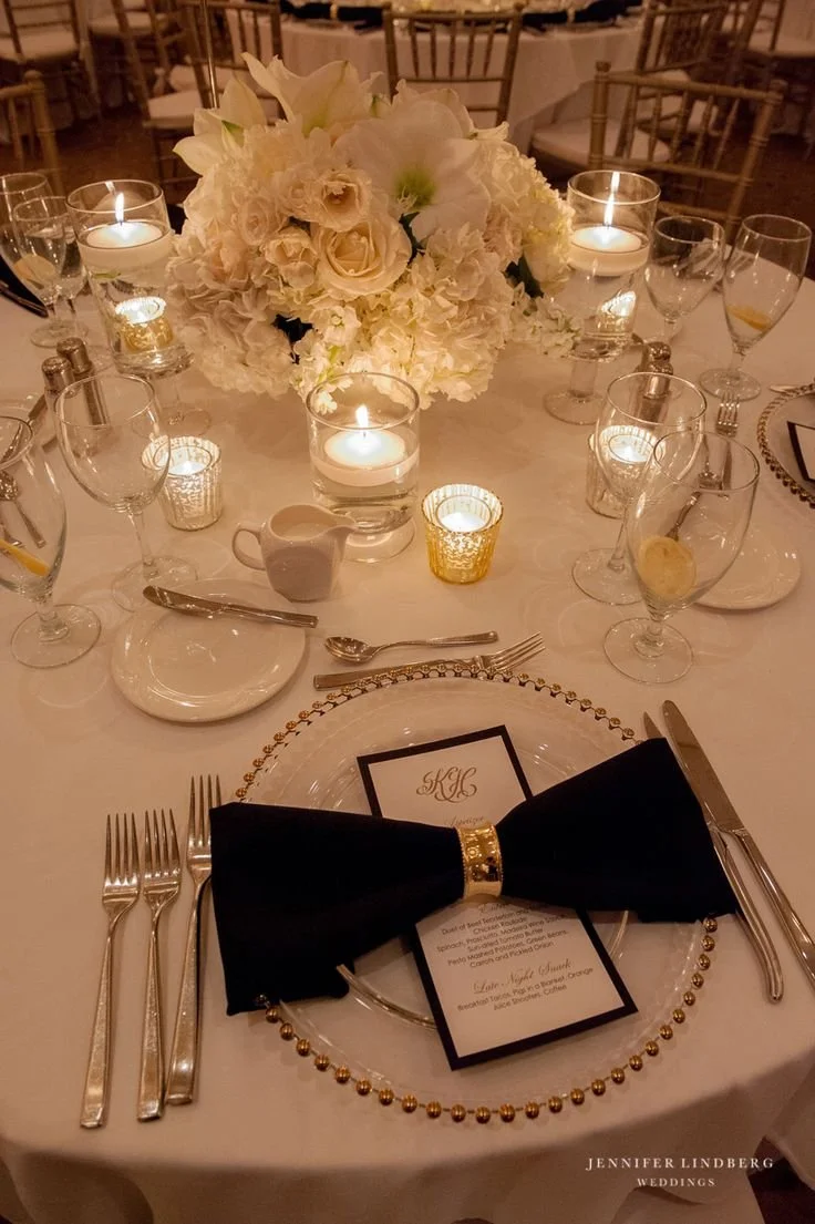 Elegant wedding reception table with white floral centerpiece, lit candles, and formal place settings, including a black bow tie on a plate with a menu card.