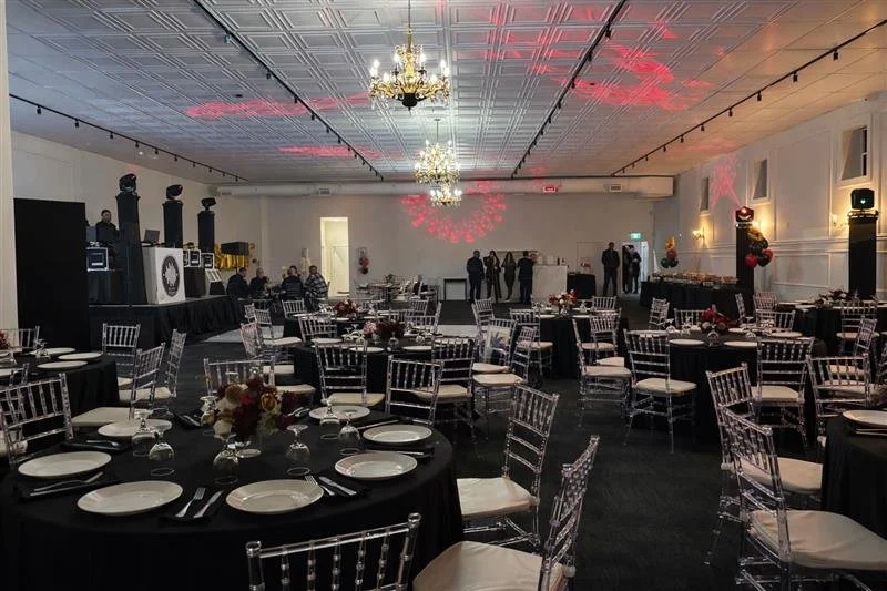 Event hall set up for a celebration with round tables, black tablecloths, white chairs, floral centerpieces, and lighting effects on the ceiling.
