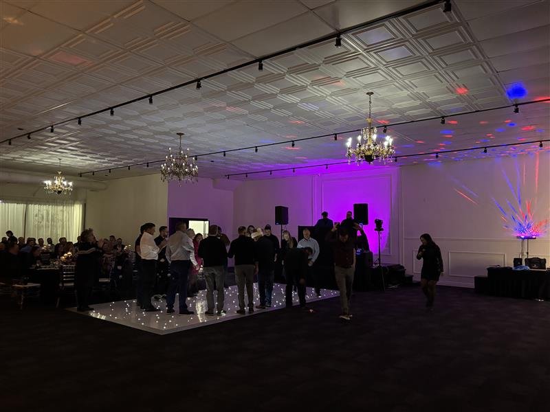 People gathered on a dance floor at a formal event with chandeliers, purple lighting, and a DJ setup, while guests sit at tables in a decorated banquet hall.