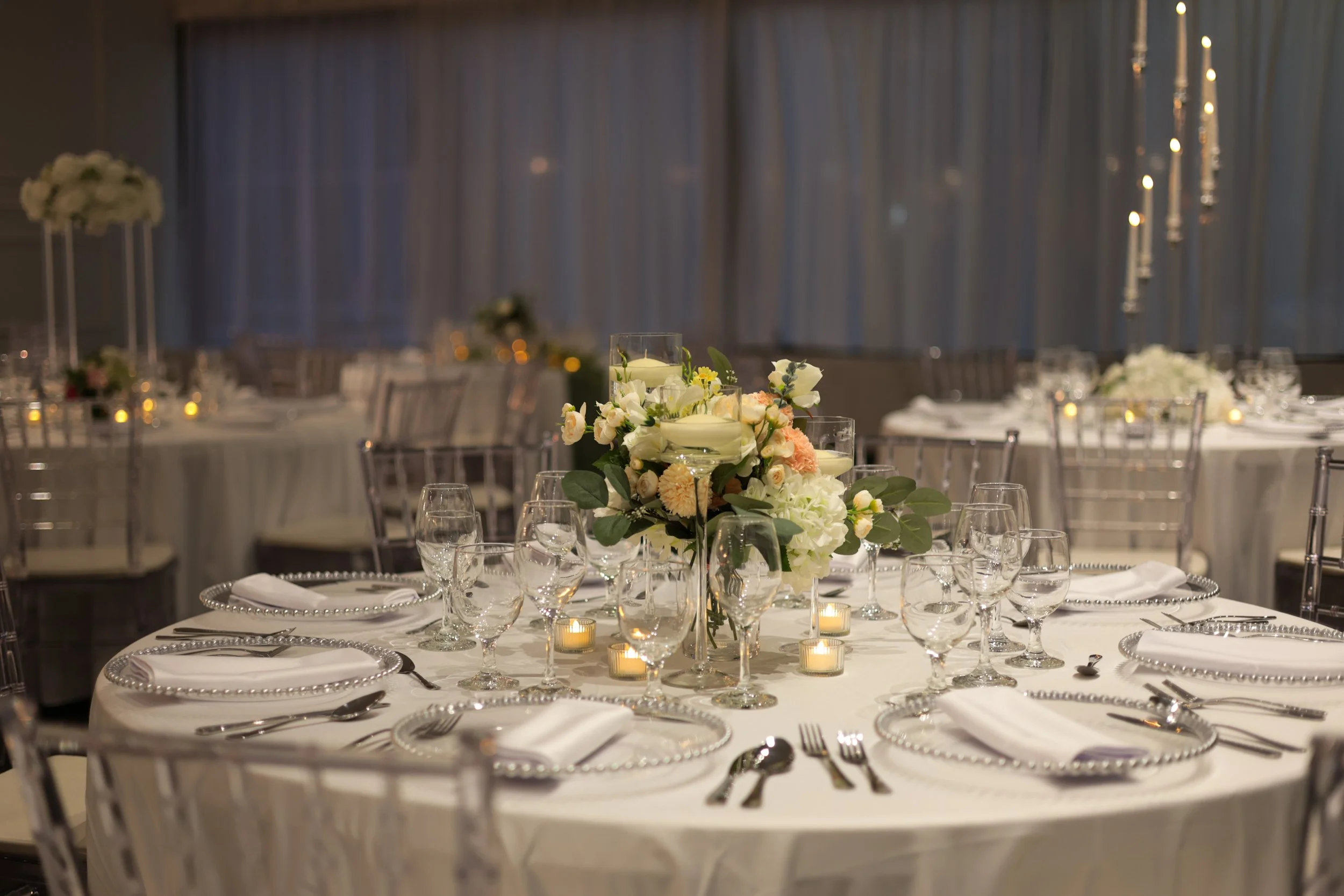 Elegant banquet table set with white tablecloth, floral centerpiece, multiple wine glasses, and lit candles in a decorated event hall.