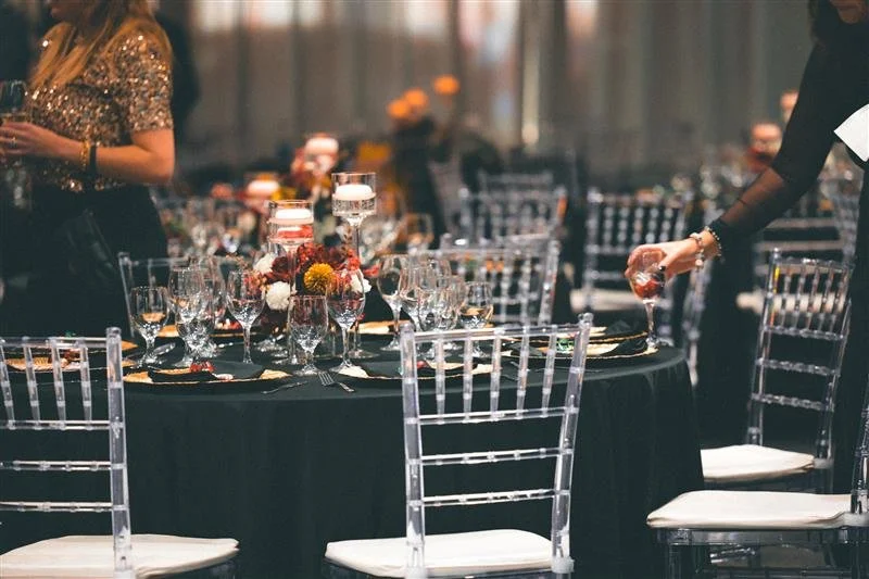 Elegant event setup with a round table, black tablecloth, floral centerpiece, glassware, and empty chairs, with people preparing the table.