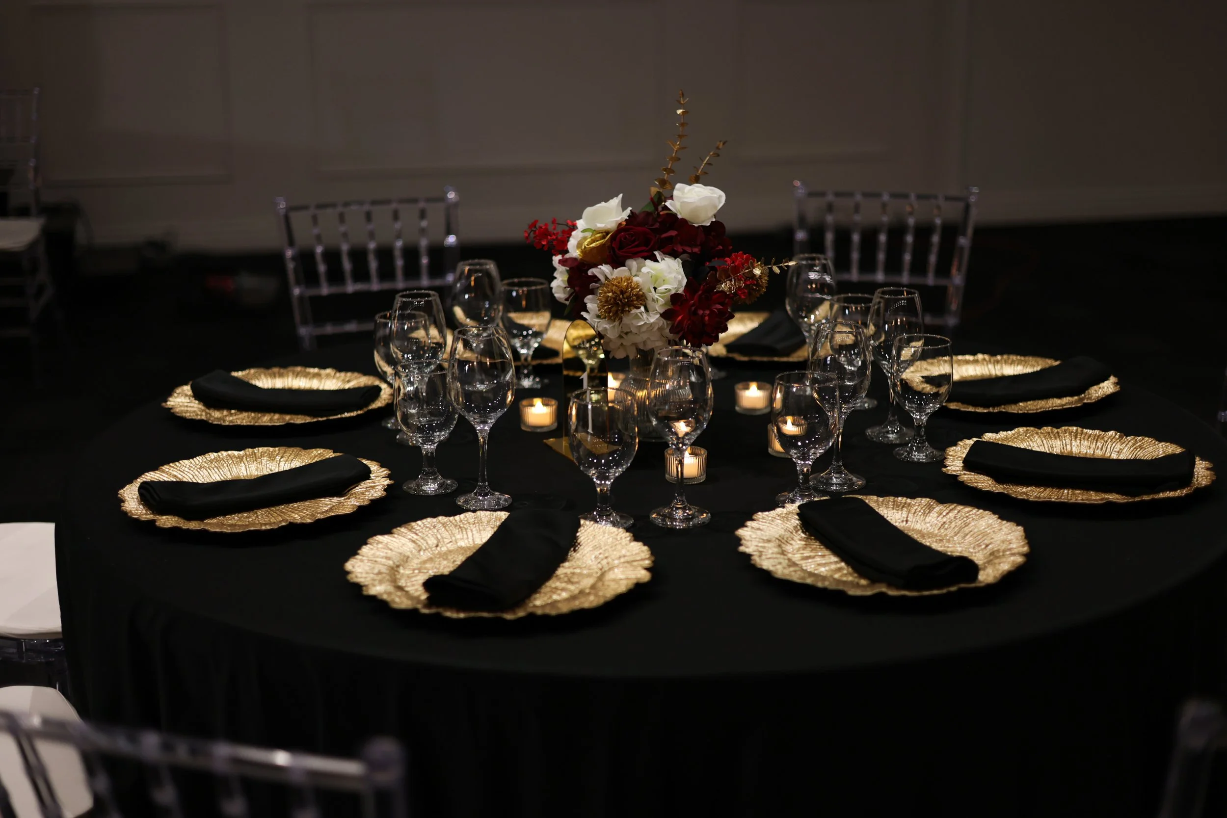 A round dining table set for a formal event with black cloth covering, gold chargers, black napkins, and glassware. Centerpiece features a large bouquet of flowers in white, red, and gold, surrounded by small candles.