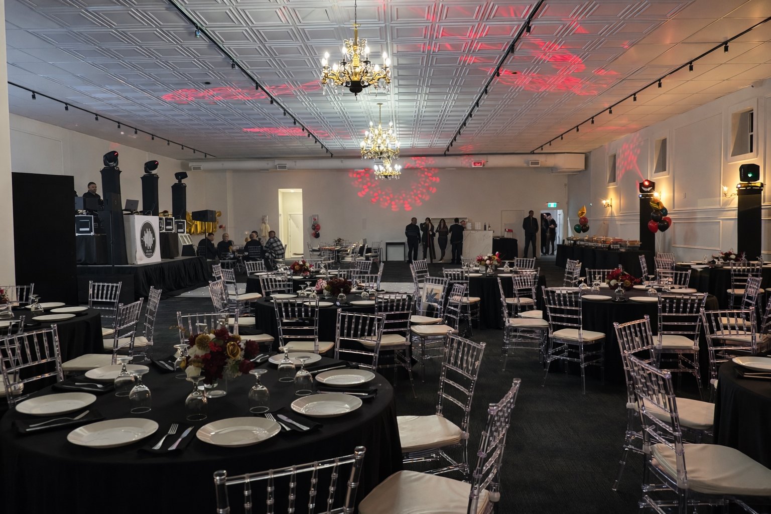 Banquet hall decorated for an event with black tablecloths, white plates, and floral centerpieces. Transparent chairs surround the tables, and a DJ booth with speakers is set up on a stage to the left. Colorful balloons adorn the right side, and several people are standing near the back of the room. Red and white lighting creates a festive atmosphere.