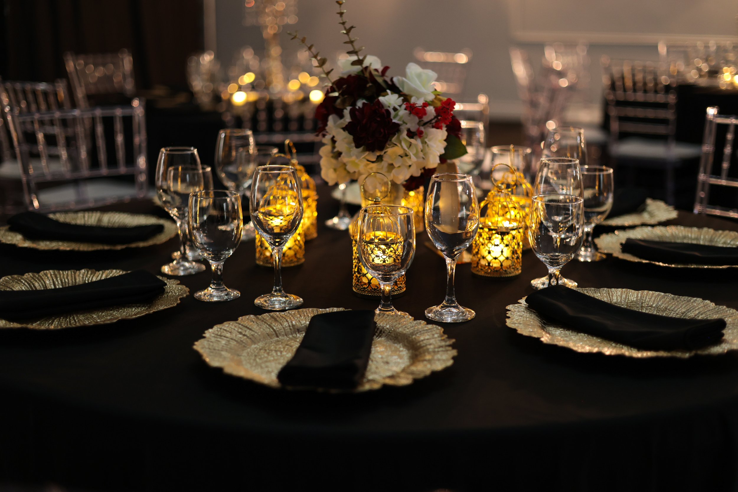 Elegant dining table set with black napkins, gold chargers, and clear wine glasses. Centerpiece of white and deep red flowers surrounded by lit gold lattice lanterns creates a lavish ambiance for a formal event. Wedding event decoration ideas. 