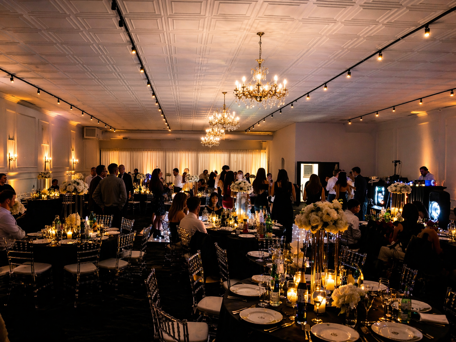 An elegant banquet hall decorated for a formal event with chandeliers, string lights, and floral centerpieces on tables with black tablecloths and clear chairs.