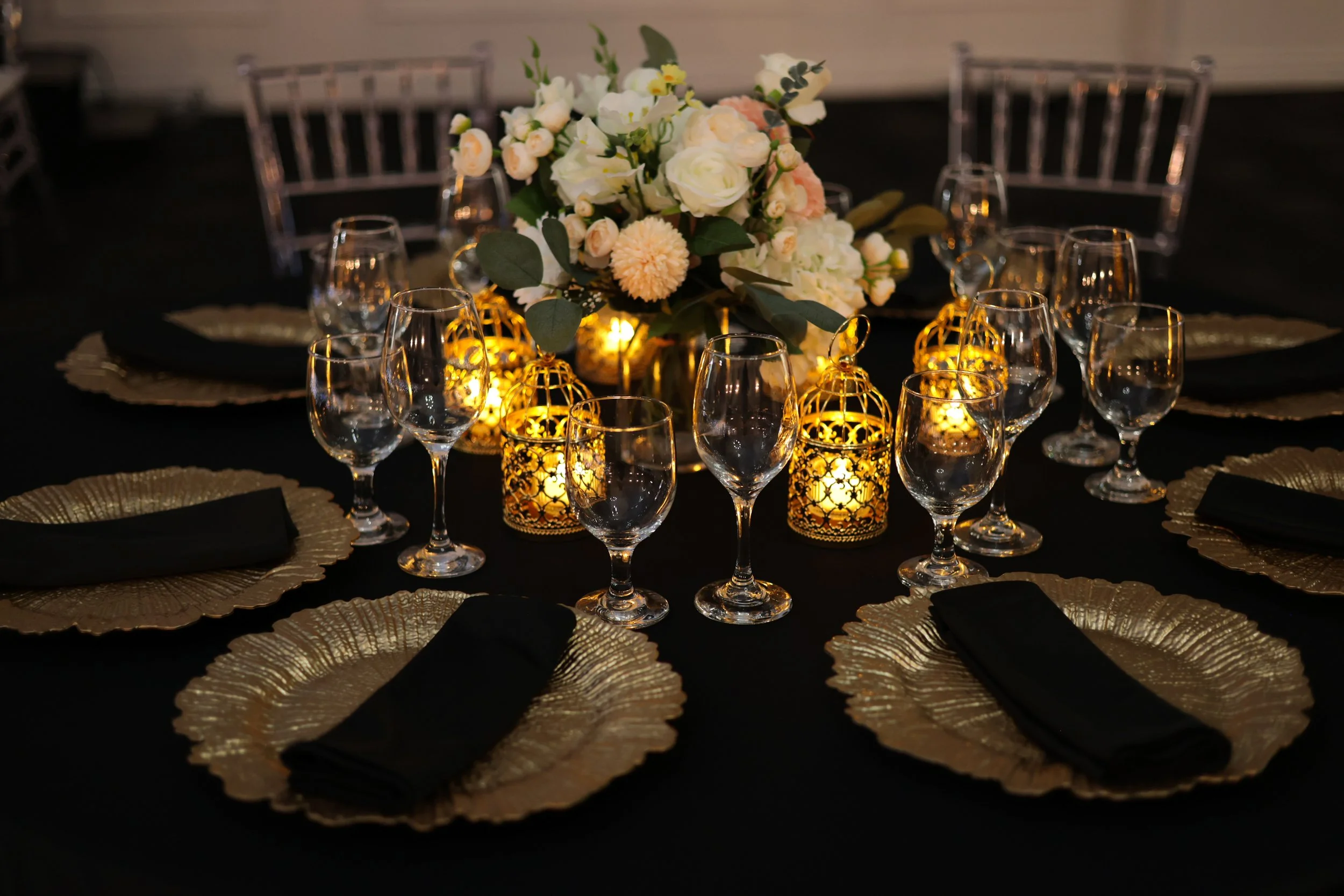 Elegant dining table setup with gold chargers, black napkins, empty wine glasses, a floral centerpiece with white and blush flowers, and decorative candle lanterns with yellow lights.