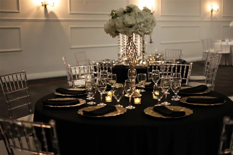 Elegant banquet table with a black tablecloth, set with gold chargers, black napkins, and glassware. Centerpiece features white flowers and hanging crystal decorations, with small candles around it.
