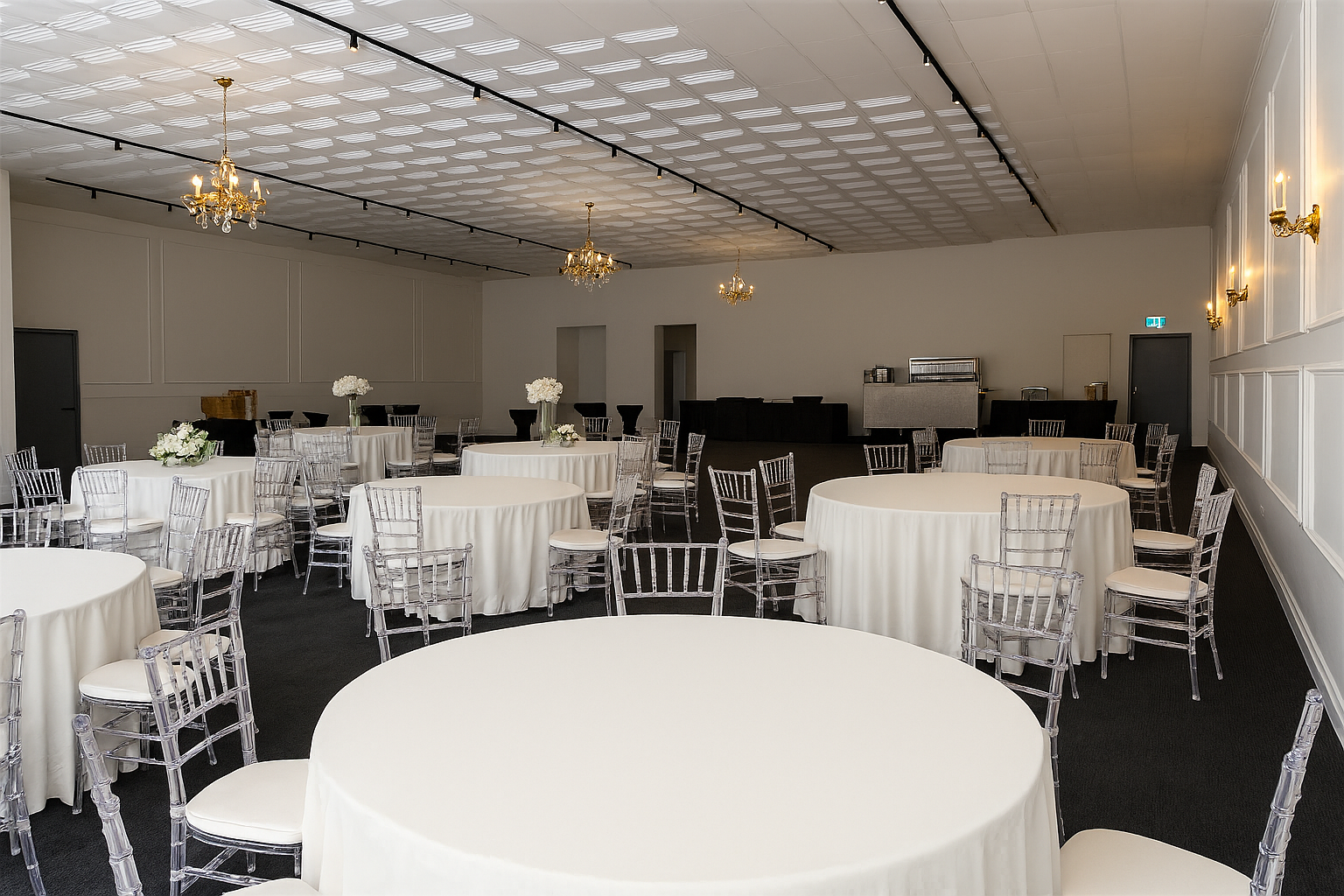 A banquet hall with white round tables covered in white tablecloths and clear acrylic chairs. The room has elegant chandeliers and wall sconces, with floral centerpieces on each table. The ceiling is decorated with a patterned design, and the back of