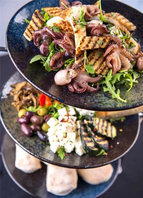 A three-tiered black serving tray holds an assortment of Mediterranean appetizers: on the top tier, grilled octopus with greens and grilled bread; on the middle tier, assorted olives, feta cheese, roasted peppers, and grilled zucchini; on the bottom, pita bread pieces.