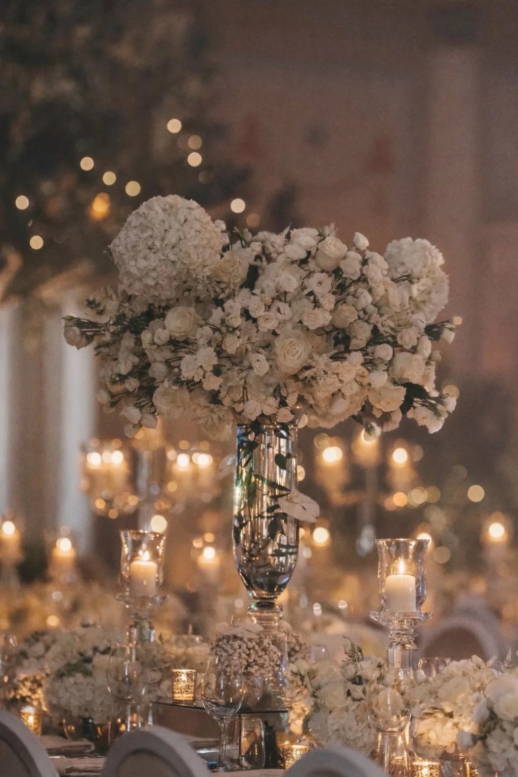 Elegant table centerpiece with white flowers in a tall crystal vase, surrounded by candles and smaller floral arrangements, at a dimly lit event or wedding reception.