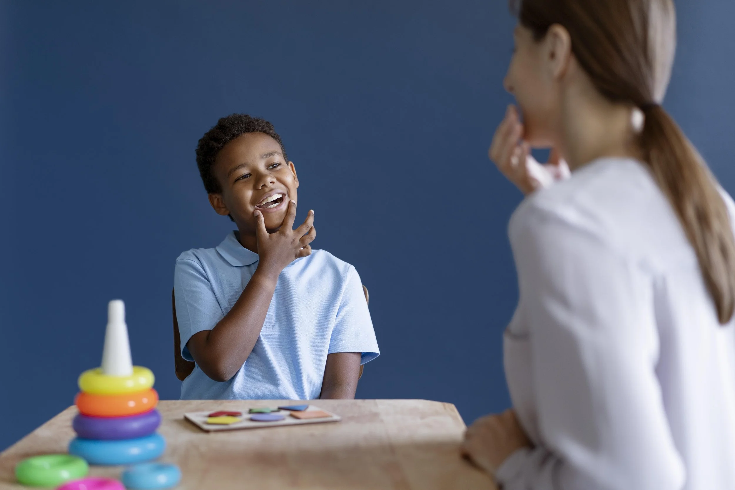 A young boy and a woman sit at a table with colorful stacking toys, engaging in a playful conversation. The boy is smiling and touching his chin, while the woman watches, smiling with her hand supporting her chin.