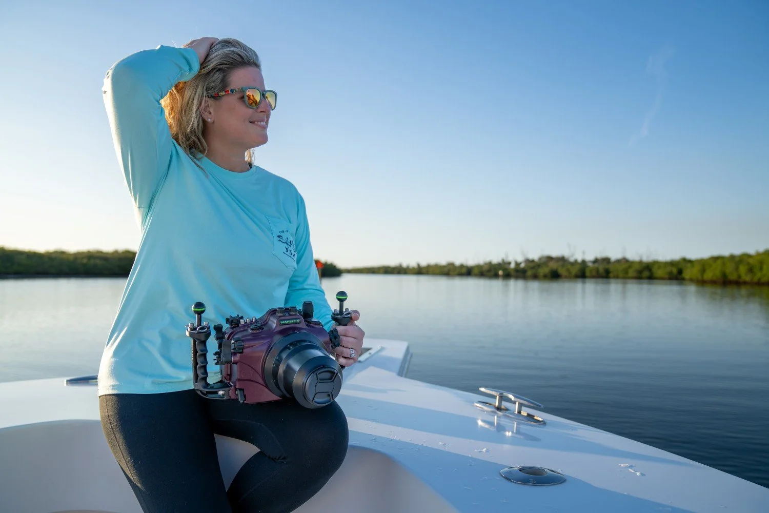 Woman wearing sunglasses and a light blue shirt sitting on a boat holding a remote control, with a body of water and trees in the background.