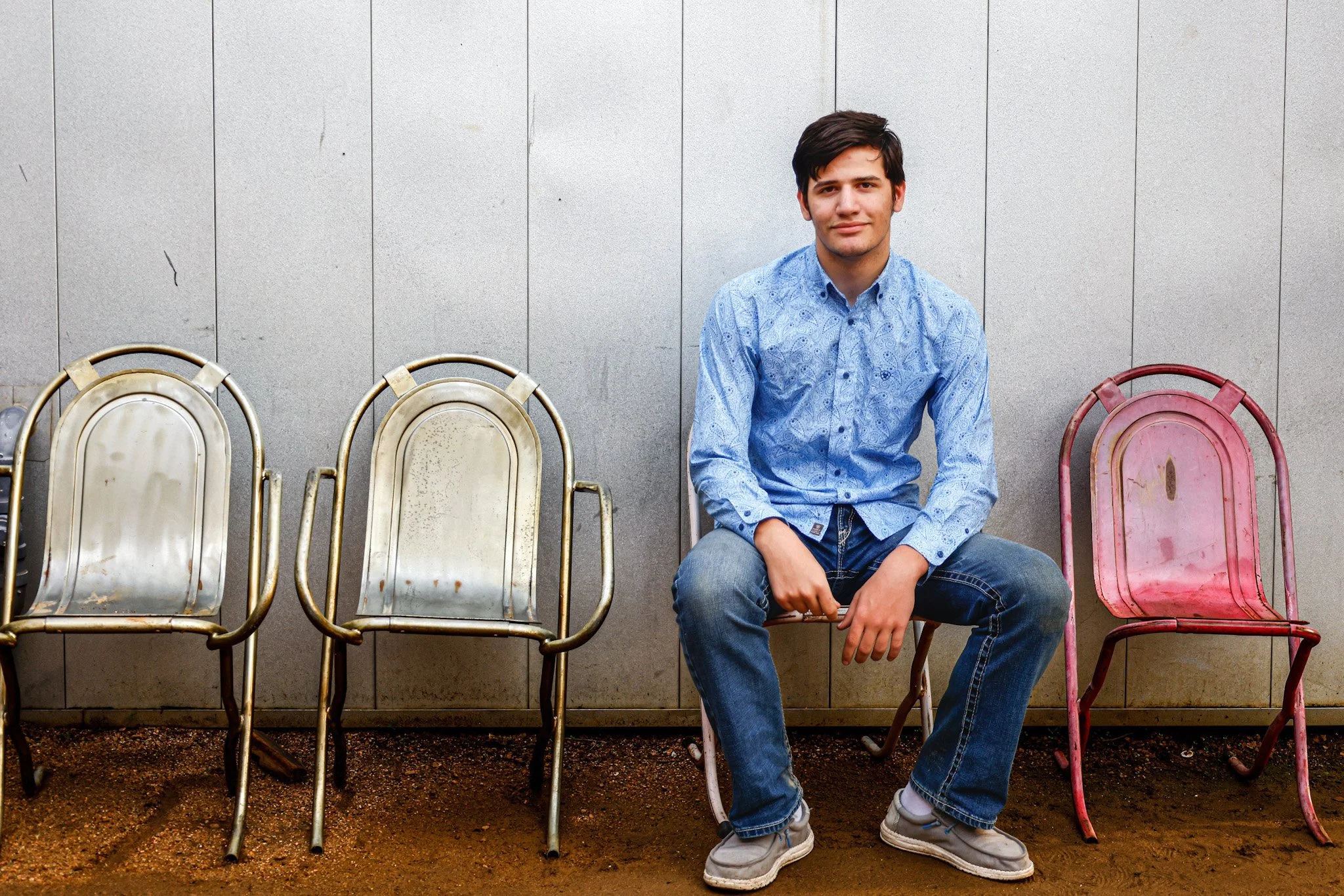 A young man sitting on a pink metal chair next to two empty chairs against a plain gray wall.