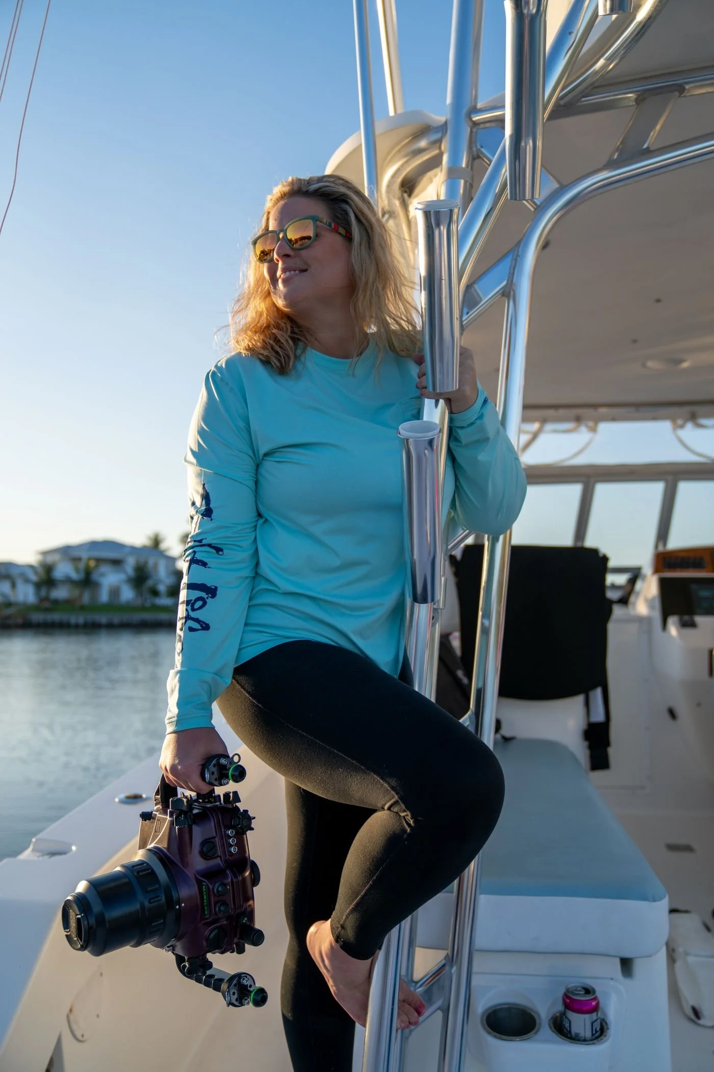 Woman with sunglasses holding a fishing rod and a camera on a boat, with water and houses in the background.