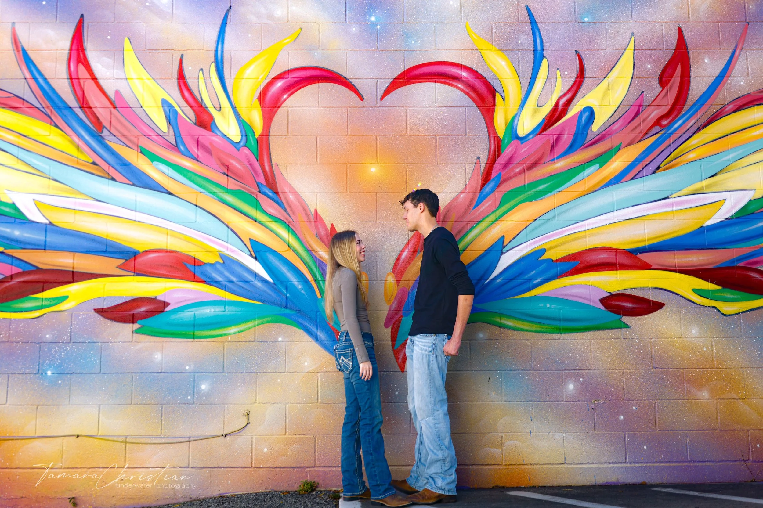A happy couple standing in front of a colorful mural of a heart with wings painted on a brick wall.
