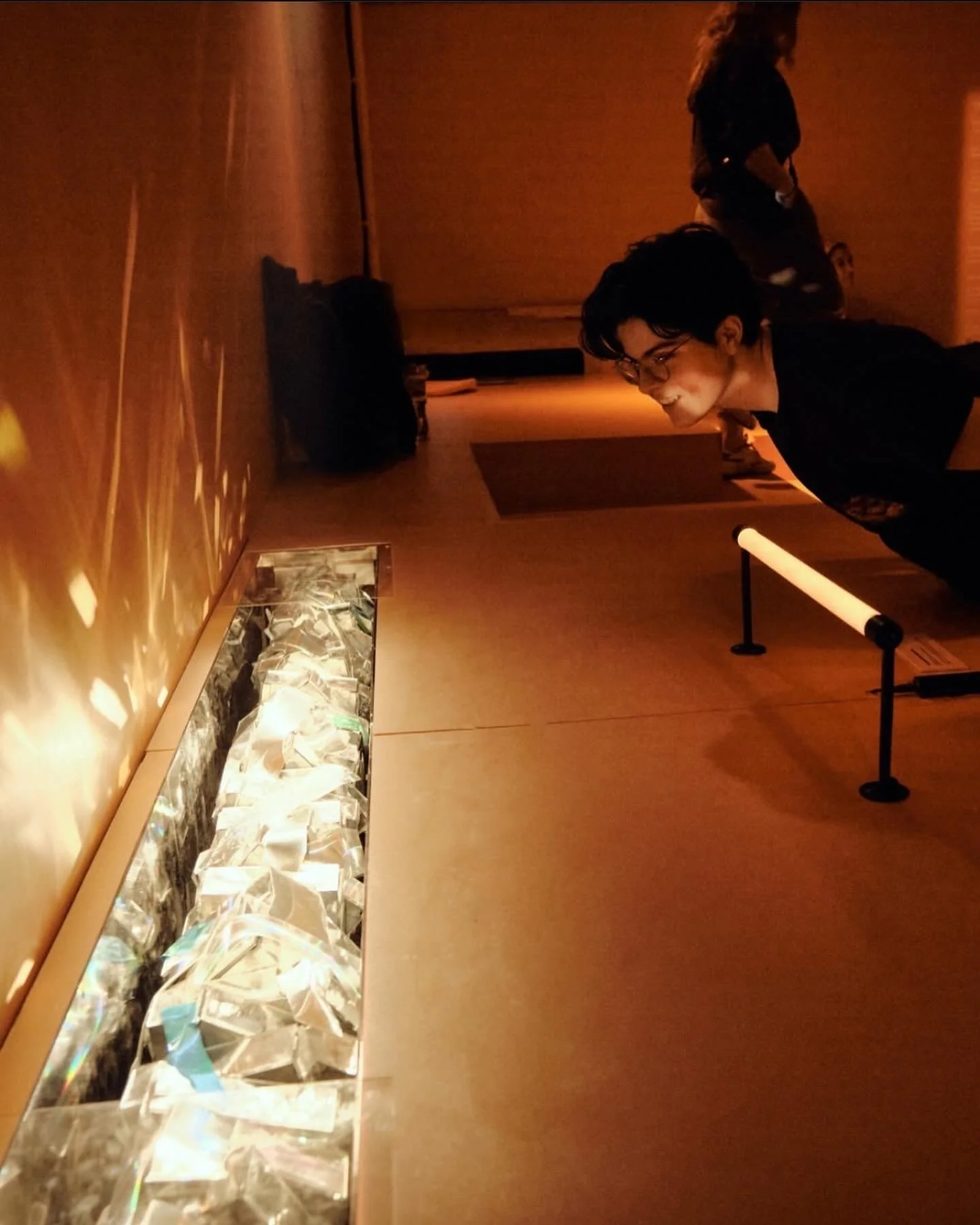 A woman and a young man admire an art installation with illuminated crumpled aluminum foil on a display case in a dimly lit museum or gallery.