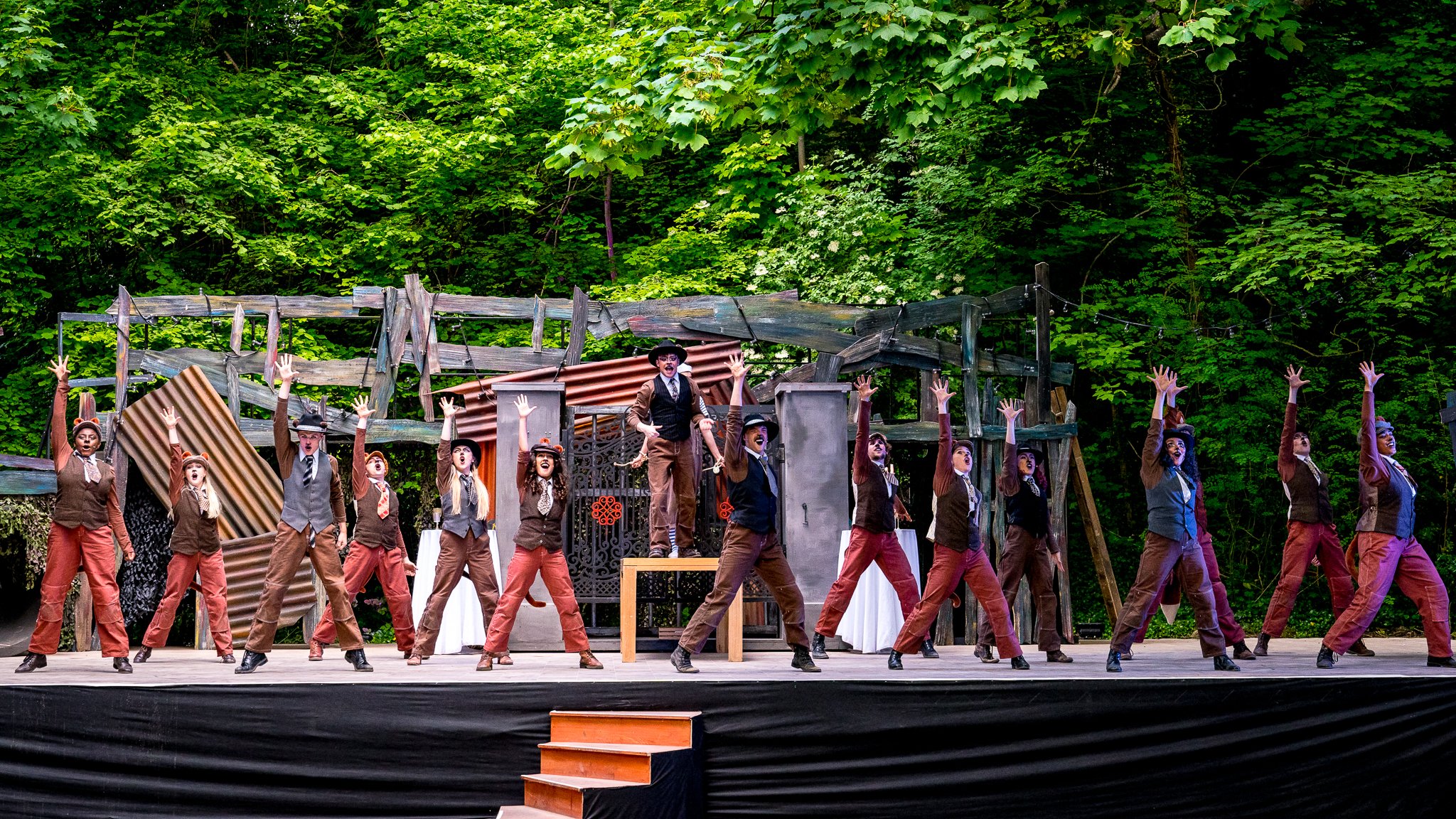Theater stage with performers in costumes, performing a musical dance routine outdoors, with a backdrop of green trees and rustic stage set pieces.