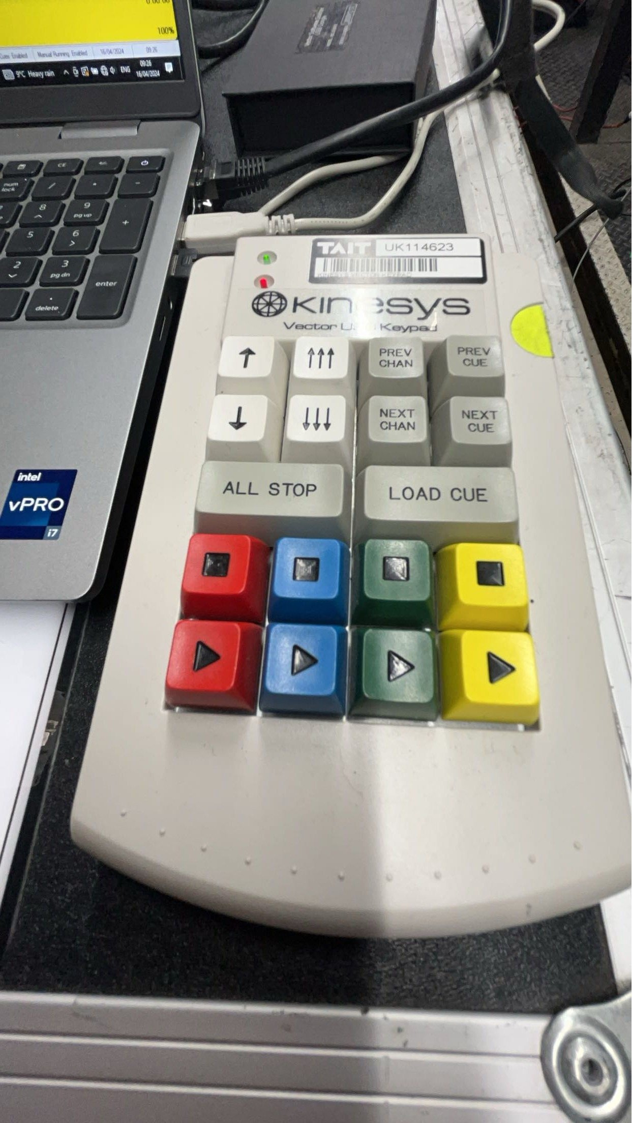 A light gray DJ controller with colored buttons (red, blue, green, yellow) on a table next to a laptop keyboard.