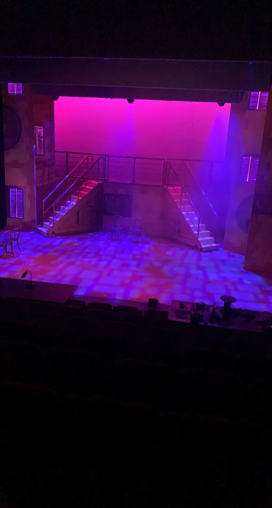 Empty theater stage with colorful lighting, two staircases, and several chairs.