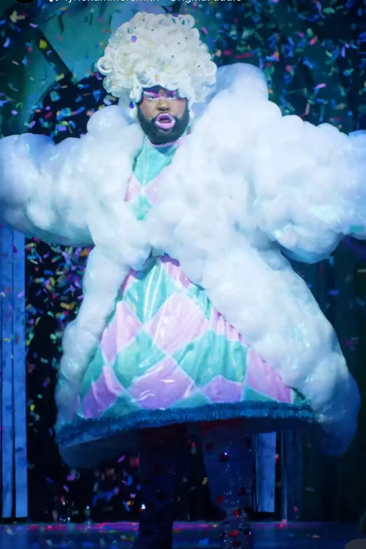 Person dressed in colorful, theatrical costume with a fluffy, cloud-like coat and pastel diamond pattern dress, wearing pink glasses and sporting a curly blonde wig, surrounded by confetti.