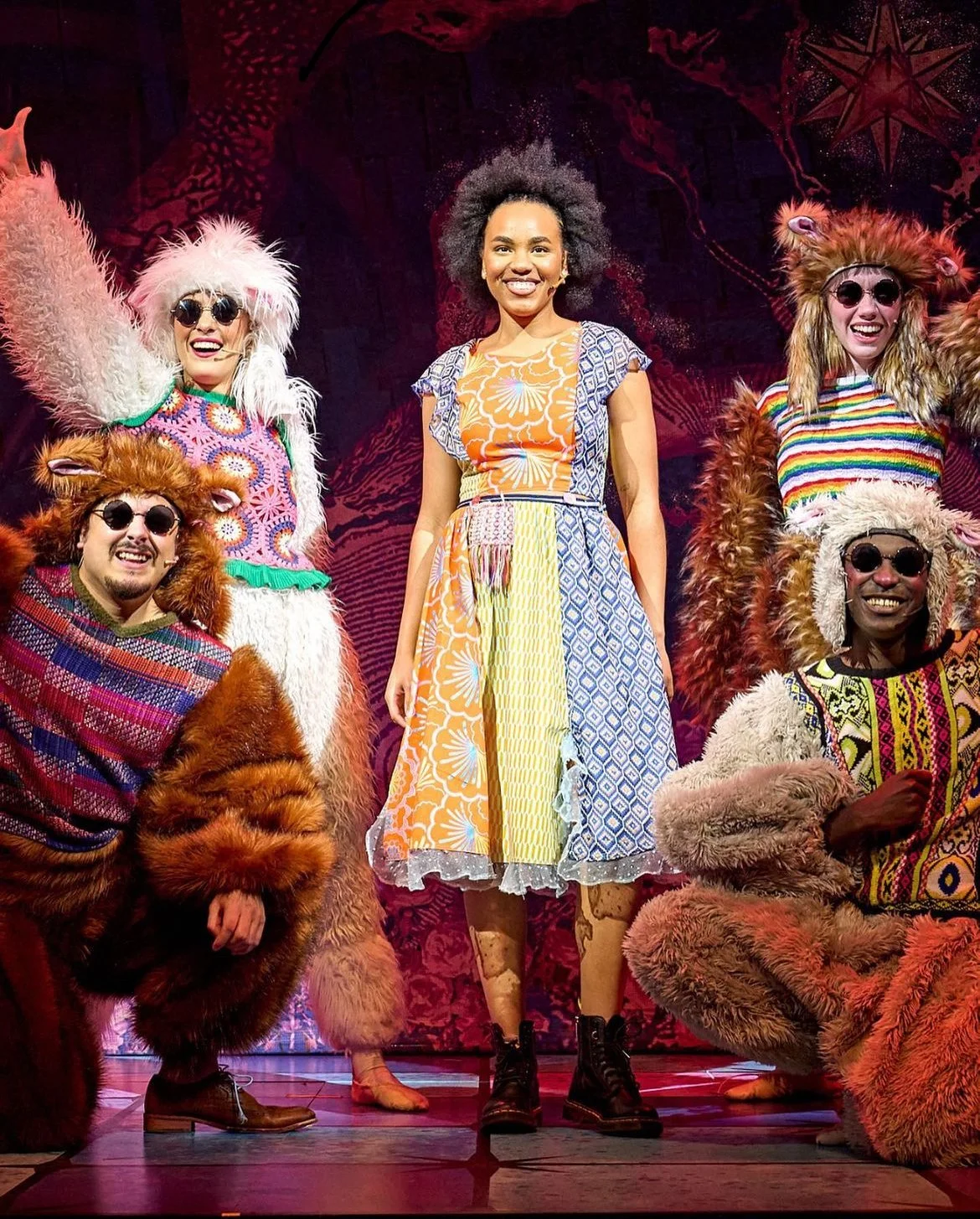 Group of four performers on stage, three in colorful furry costumes with animal ears and sunglasses, and one woman in a multicolored dress, all smiling.