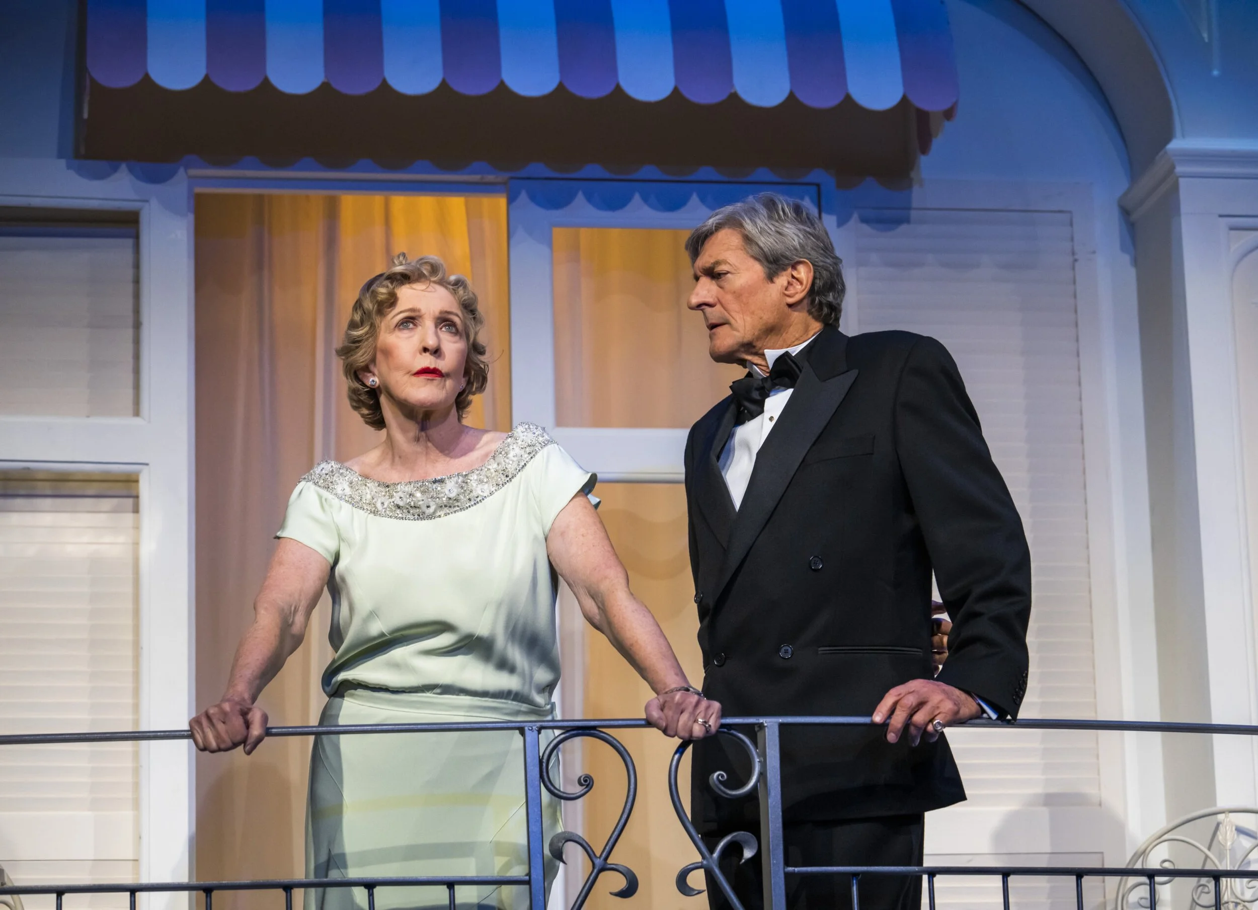 Older woman and man in tuxedo having a serious conversation on a balcony stage with a house set in the background.