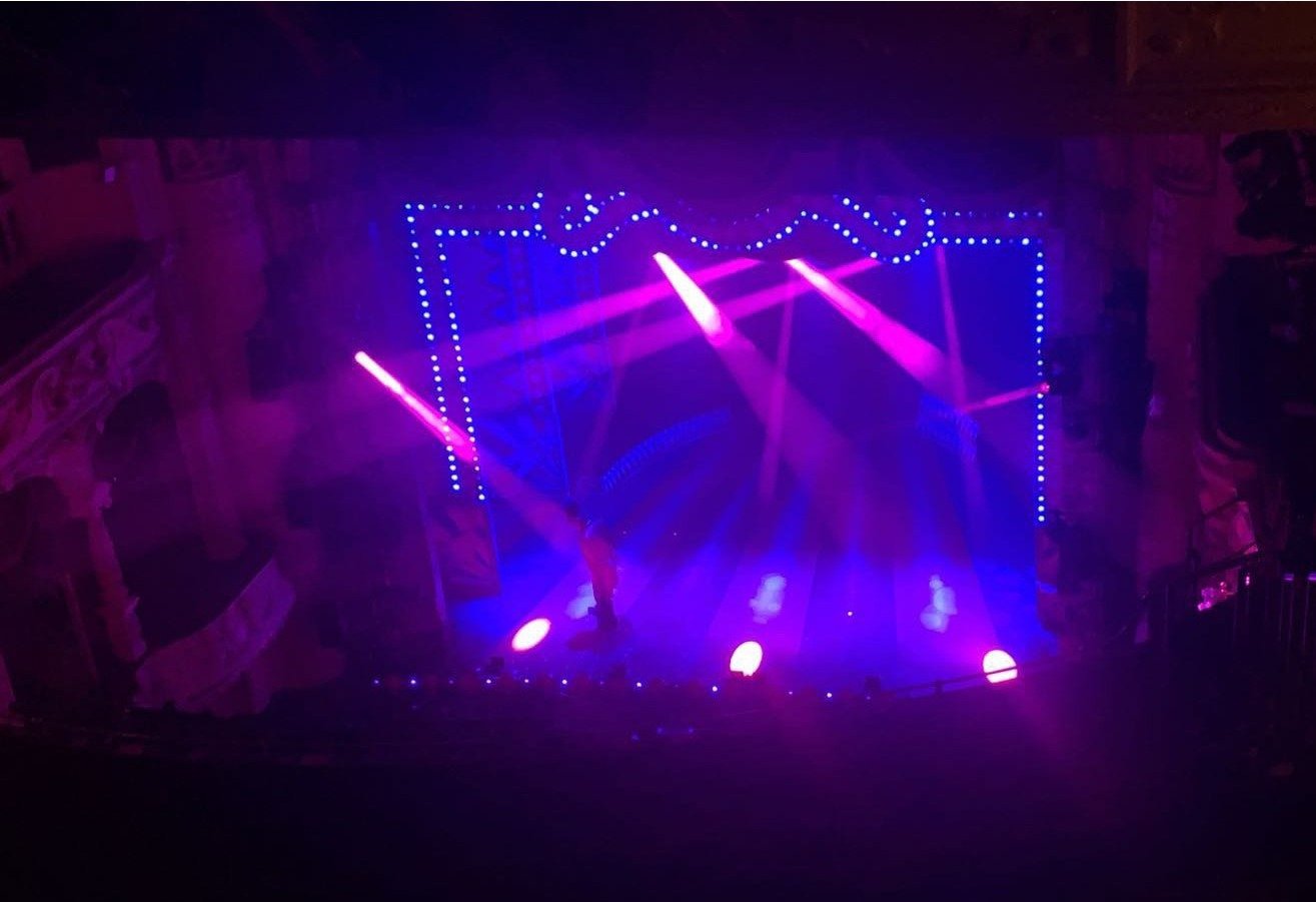 A stage with purple and pink lighting, dotted blue light borders, and spotlights shining down in a dark theater or concert hall.
