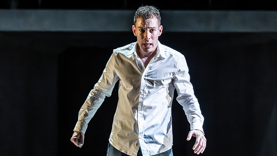 A man wearing a white button-up shirt appears to be performing on a dark stage, with an intense facial expression and body language.