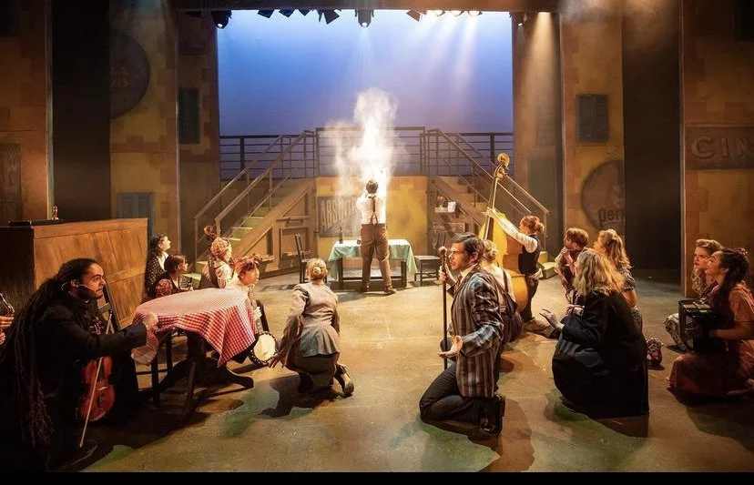 Theatrical stage performance with actors kneeling or sitting on the floor, musicians playing instruments, and a performer standing in the center with smoke rising from a device, all in a setting resembling a rustic outdoor scene.
