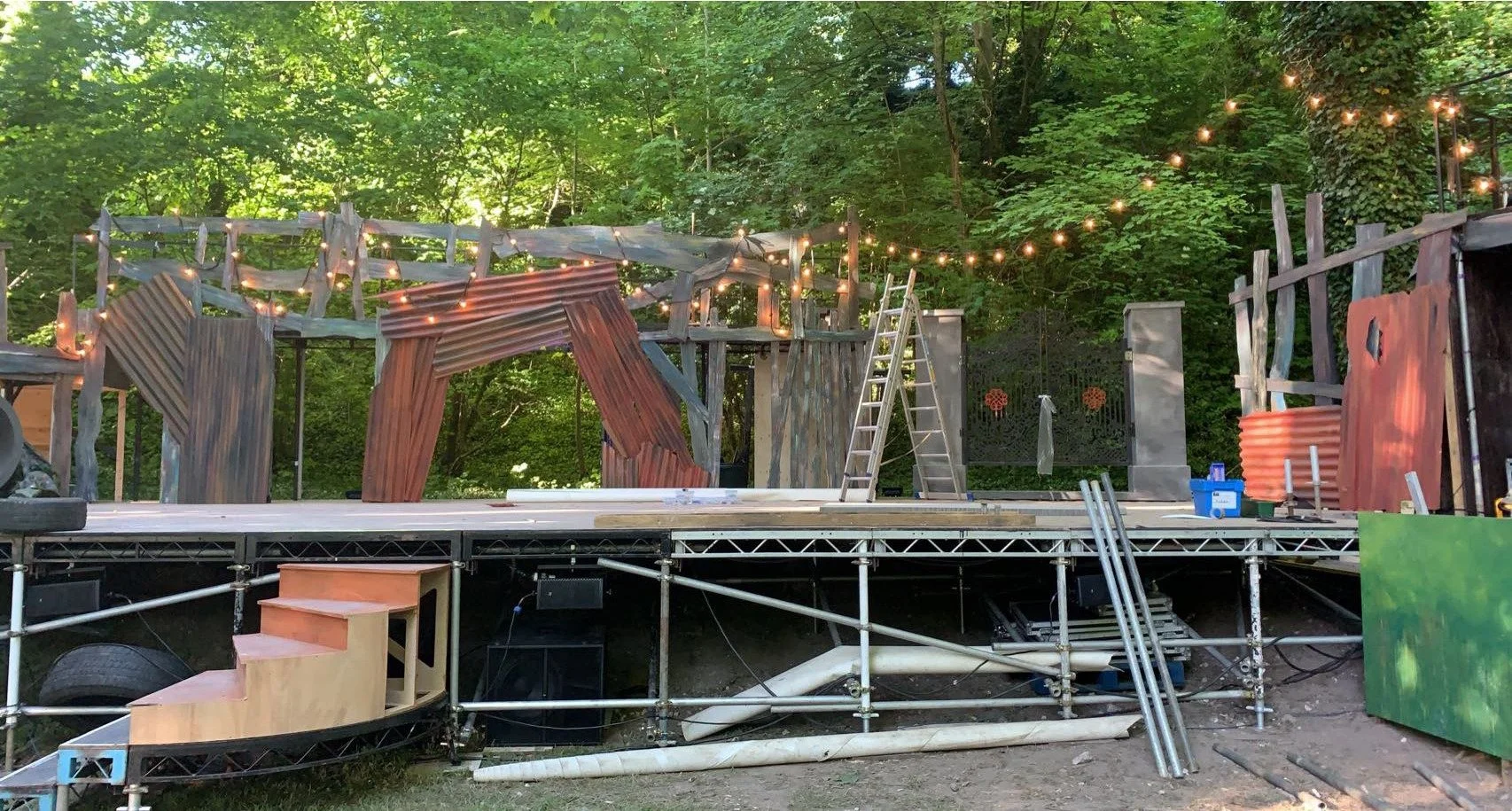 An outdoor stage under construction with wooden panels, ladders, and string lights, surrounded by green trees.