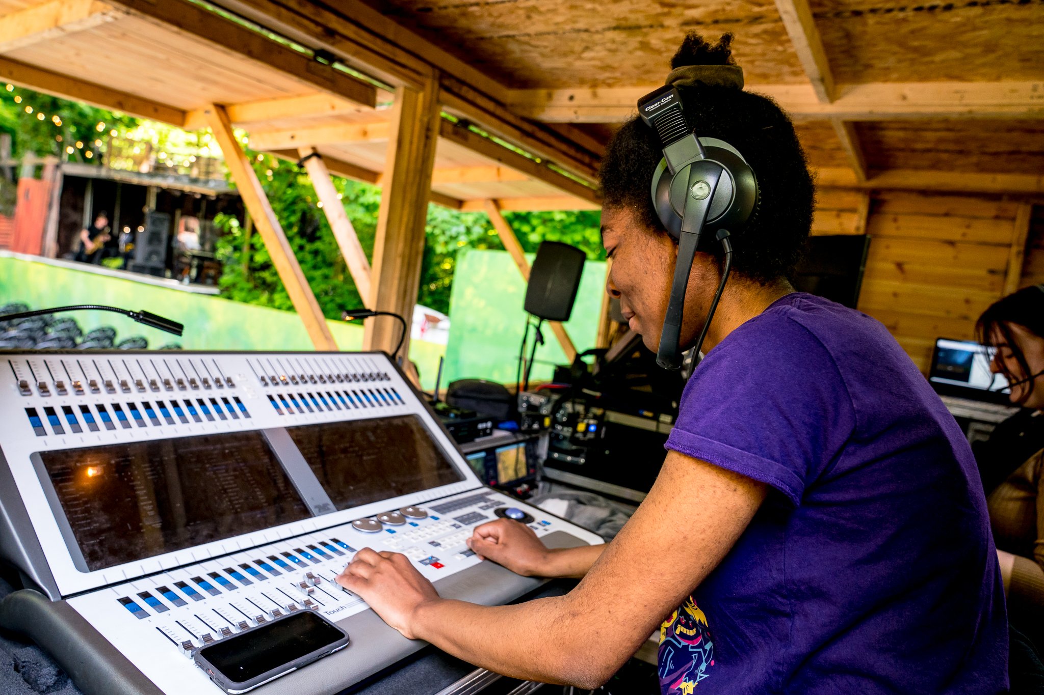 A person operating a lighting console wearing headphones, with another person working on a laptop nearby inside a wooden structure with outdoor scenery visible through the windows.