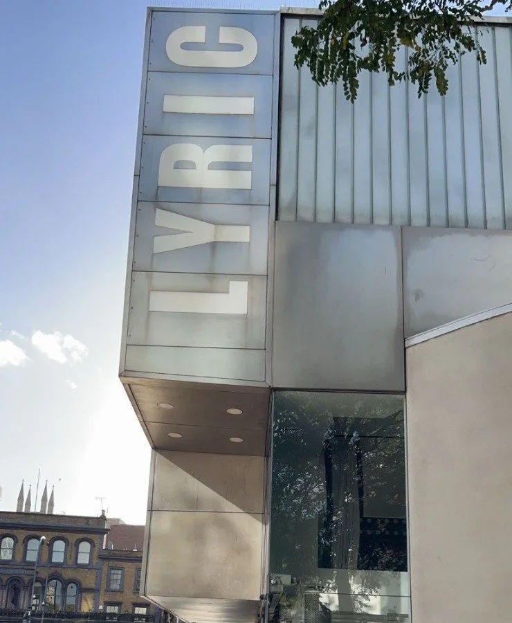 Close-up of a building with a vertical sign spelling 'LYRIC' on the corner, featuring modern design with metallic and glass elements, and some green foliage at the top right.