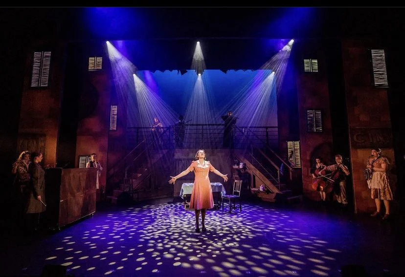 The stage of a theatrical performance with a woman in a pink dress center stage, illuminated by spotlights, surrounded by musicians and actors, with a multi-level set and dramatic lighting creating a theatrical atmosphere.