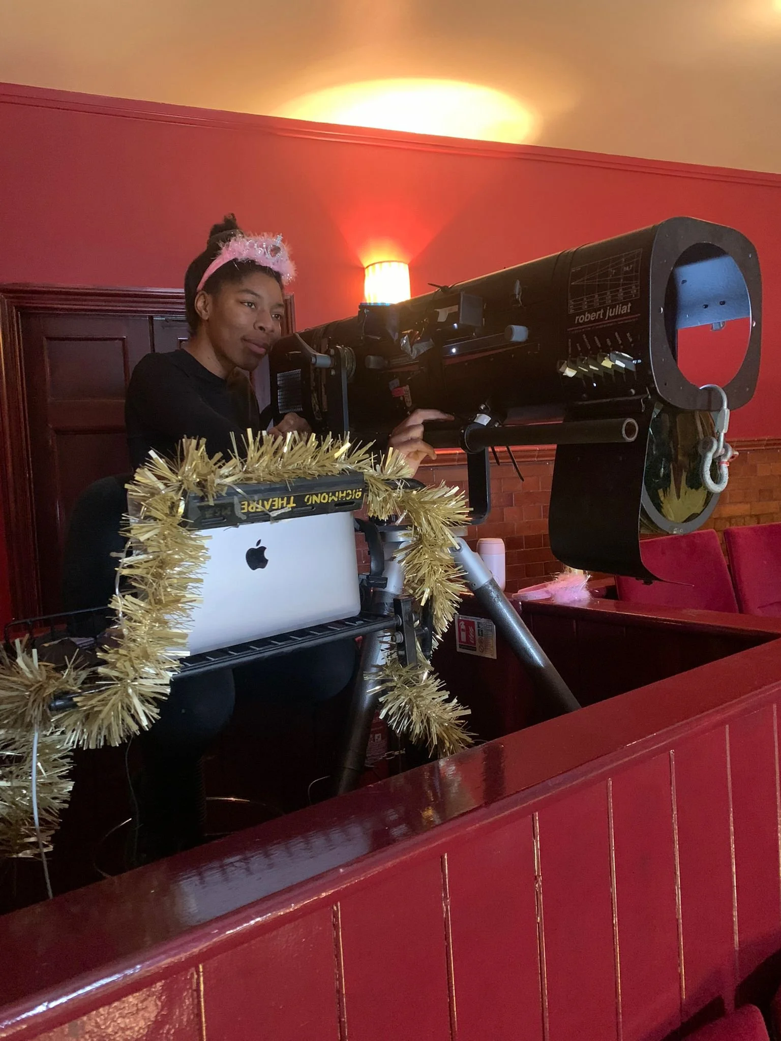 A woman, wearing a pink headband with a fluffy pom-pom, is operating an oversized telescope-like device. The setup is decorated with gold tinsel. She is inside a room with red walls, a wooden panel, and a lamp on the wall.