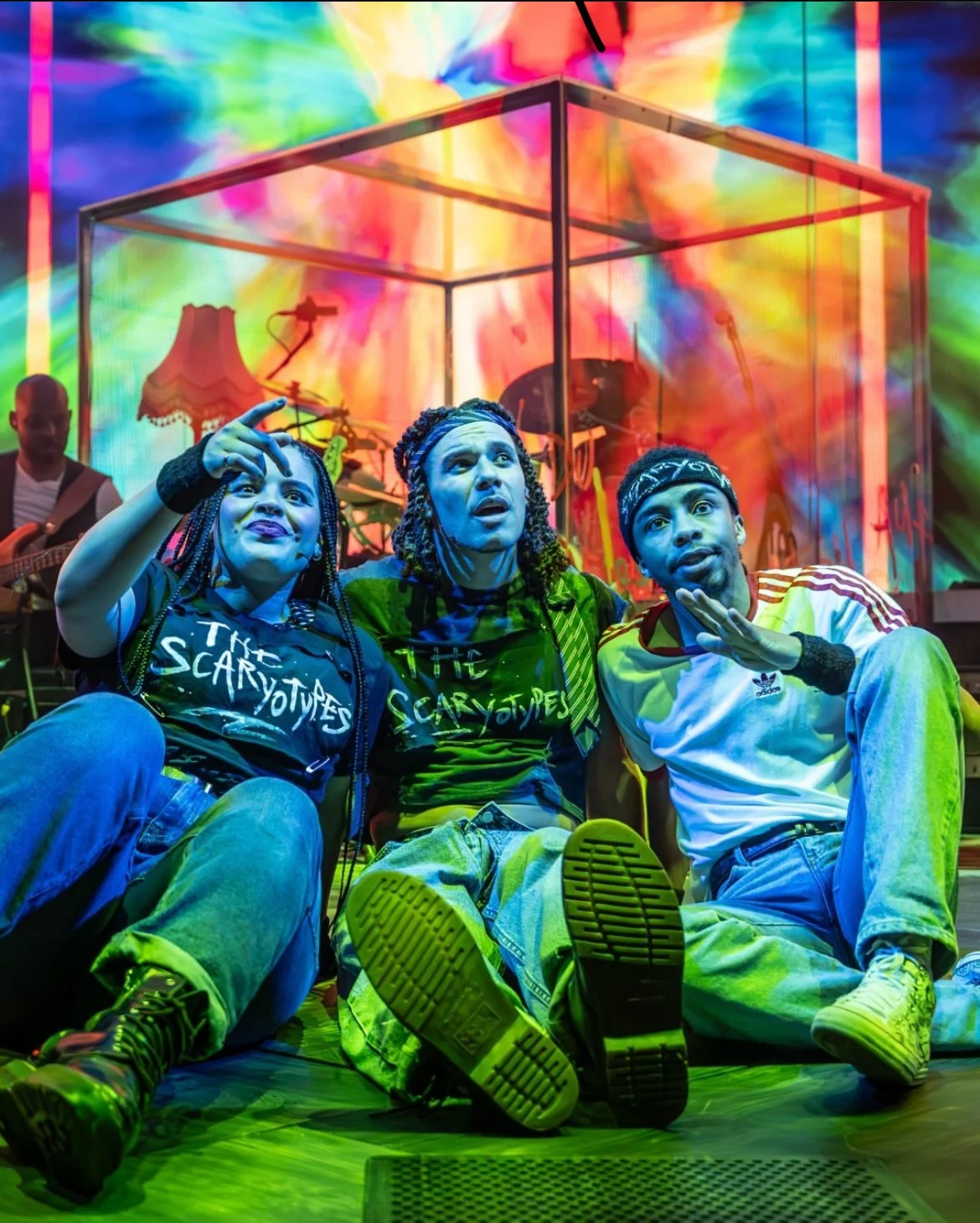 Three young people sitting on the floor in front of a colorful stage with a band playing behind a transparent box. They are making expressive faces, with the person on the left pointing toward the stage, and the person on the right with their hand ne