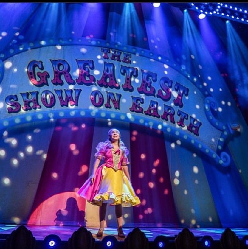 A performer on stage in colorful costume during a show titled 'The Greatest Show on Earth' with bright stage lights and decorative backdrop.