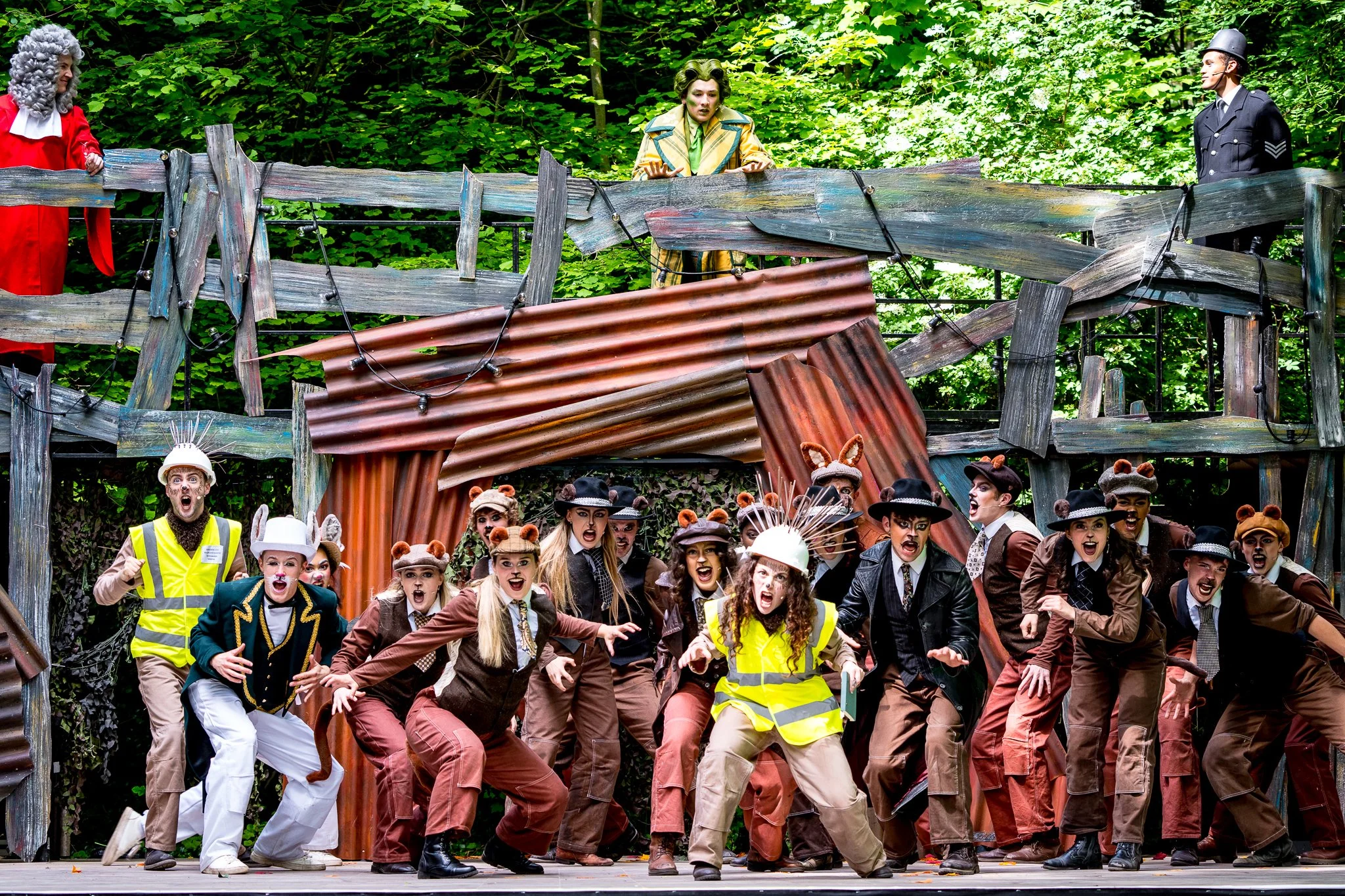 A group of actors dressed as animals, police officers, and workers perform on stage with a set resembling a forest and a rusty metal structure, depicting a scene from a play or musical.
