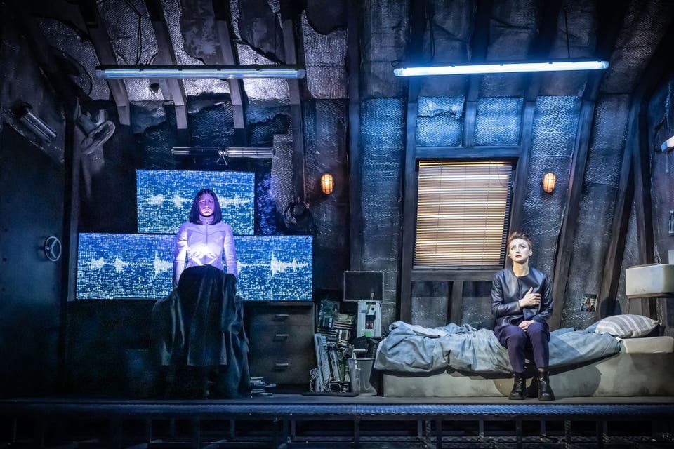 Two women are in a dimly lit room with tech equipment. One woman stands in front of multiple monitors displaying static, and the other woman sits on a bed with a distressed look.