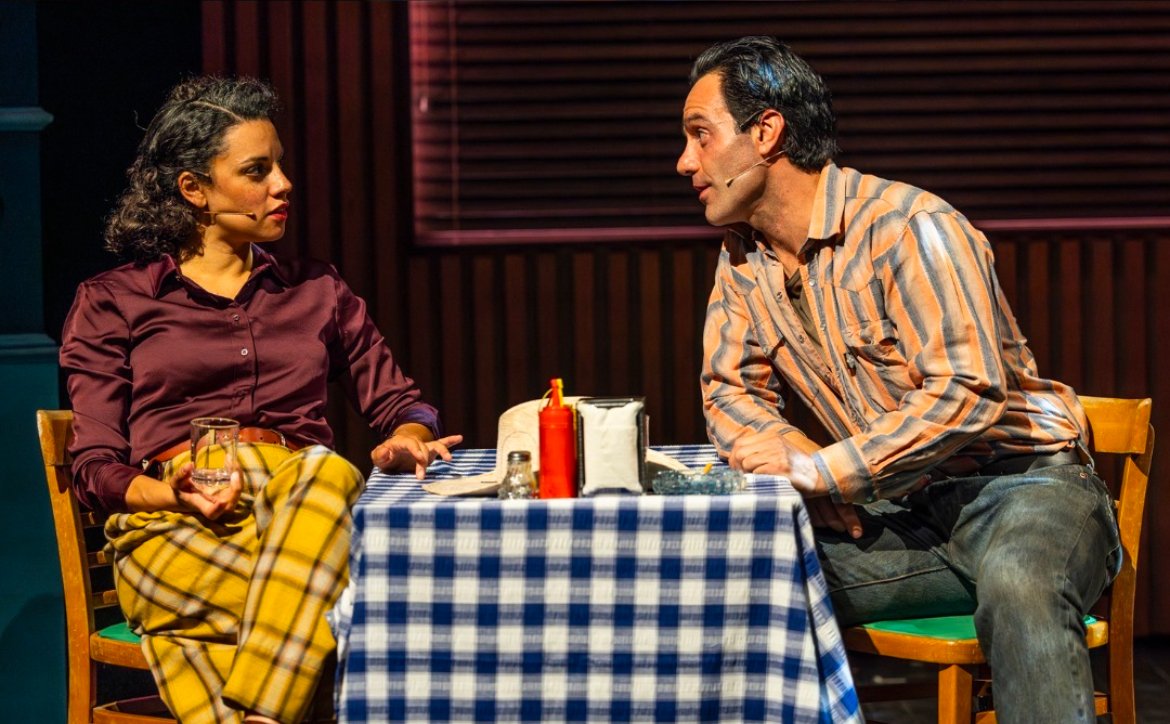 Two actors on stage engaging in a conversation at a restaurant table with checkered tablecloth, condiments, and a glass of water, during a theatrical performance.